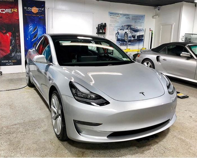 Silver Tesla sedan displayed indoors in a showroom, with a second car visible behind it.