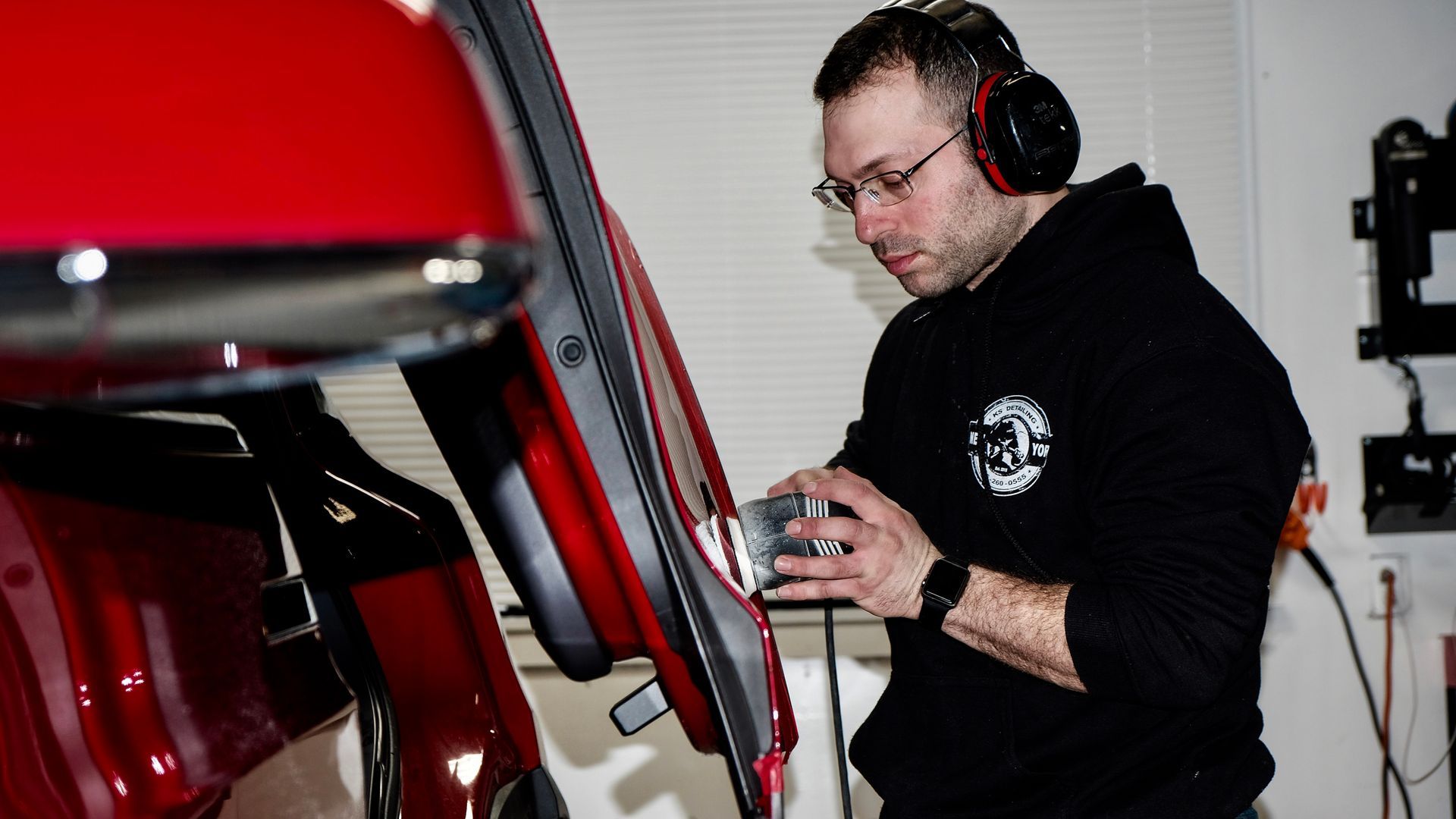Detailer polishing a red car with a buffer, wearing headphones and a black KS Detailing jacket
