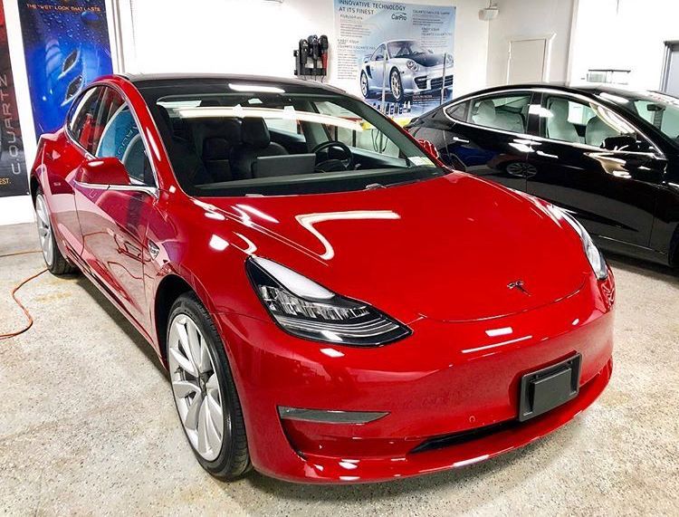 Red Tesla sedan in a showroom, with a black car parked beside it.
