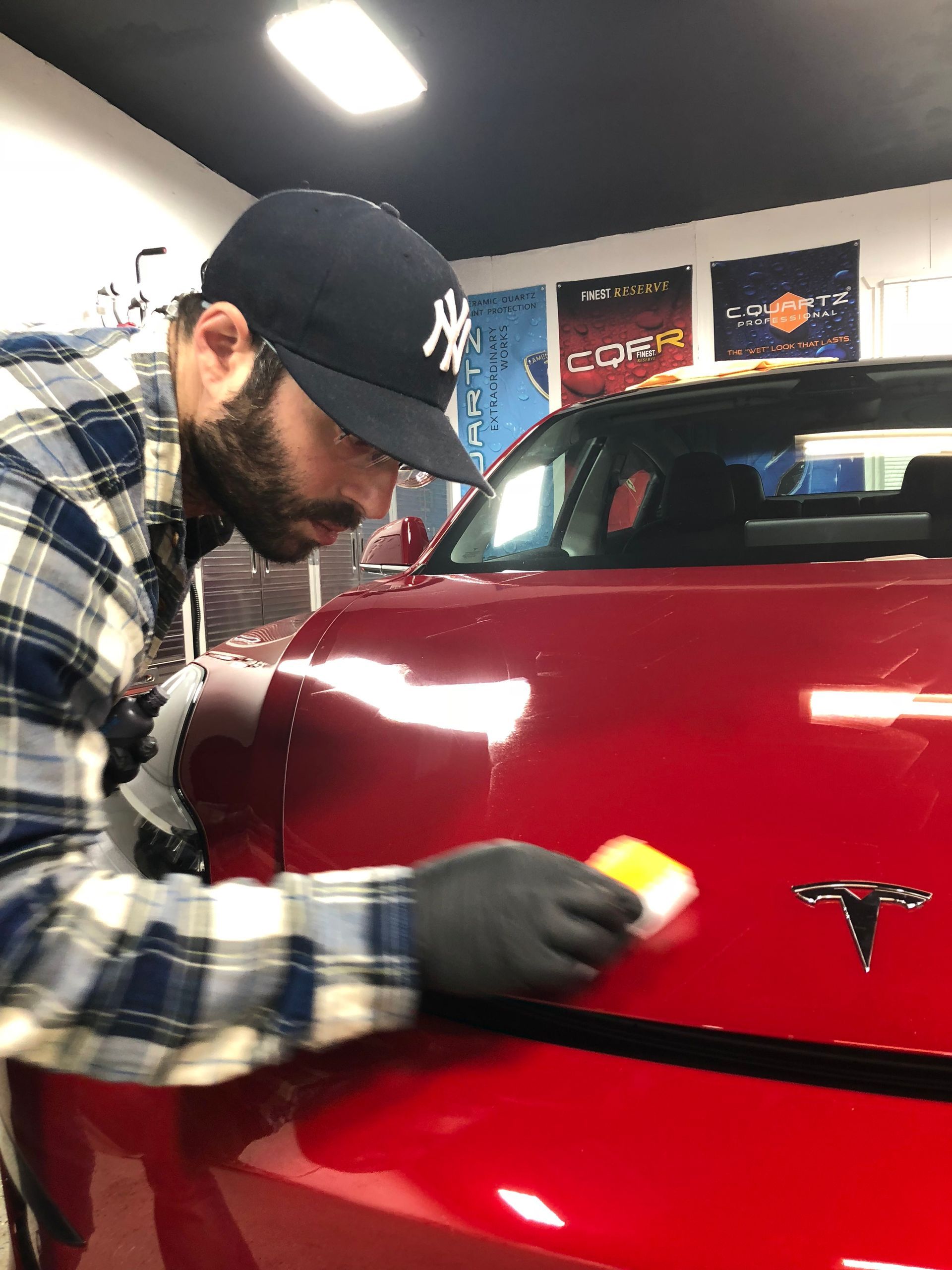 Person polishing a red Tesla hood in a garage with a sponge, wearing a cap and gloves.