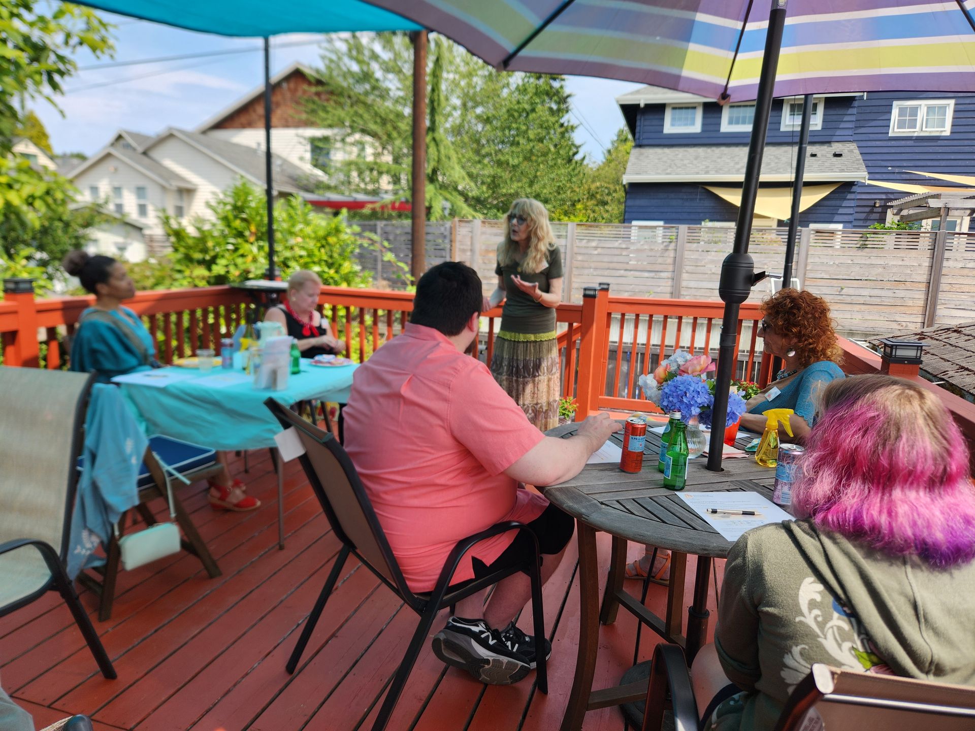 People on a deck, some seated at tables with craft supplies. A woman is standing and speaking to them under an umbrella.