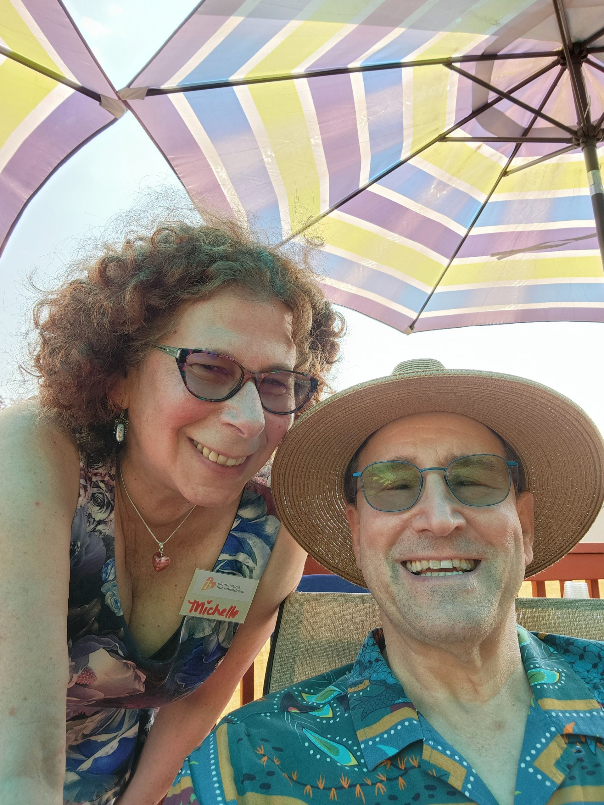 Woman and man smiling, under a striped umbrella. Outdoor setting. Woman has curly hair and glasses, and man wears a straw hat and glasses.