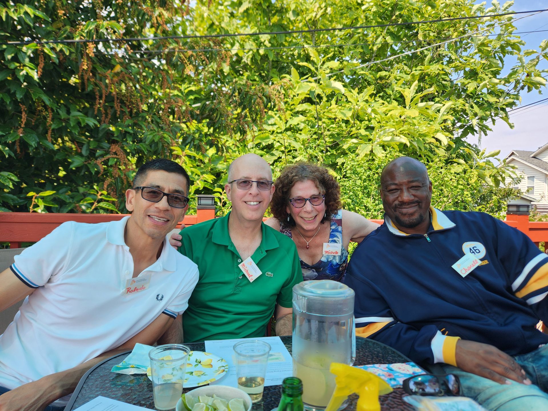 Four people seated at an outdoor table. One man wears sunglasses and a white polo. Others wear green and blue shirts. Sunny day.