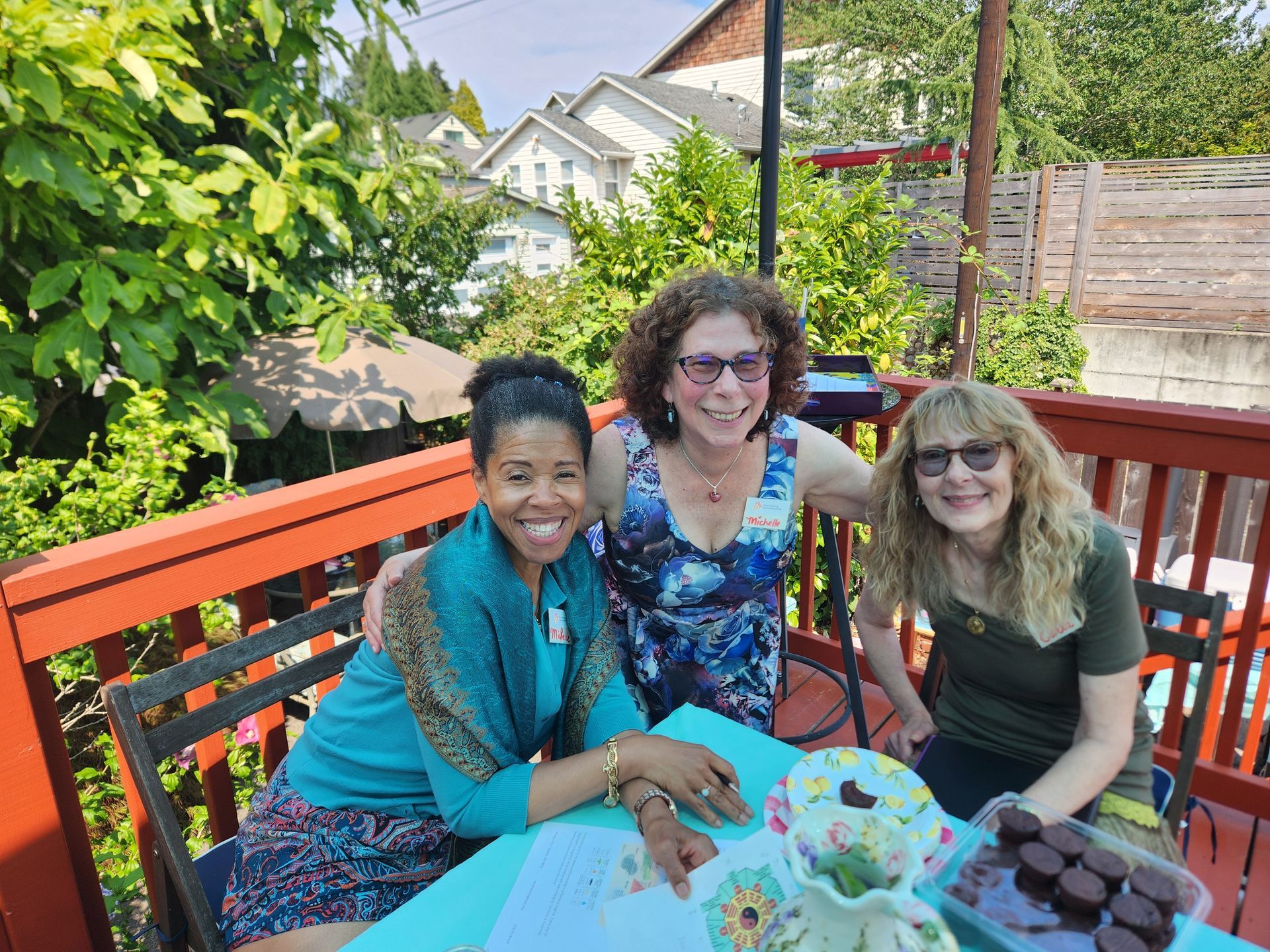 Three people smiling on a red deck, around a table with treats. Houses and trees are in the background.