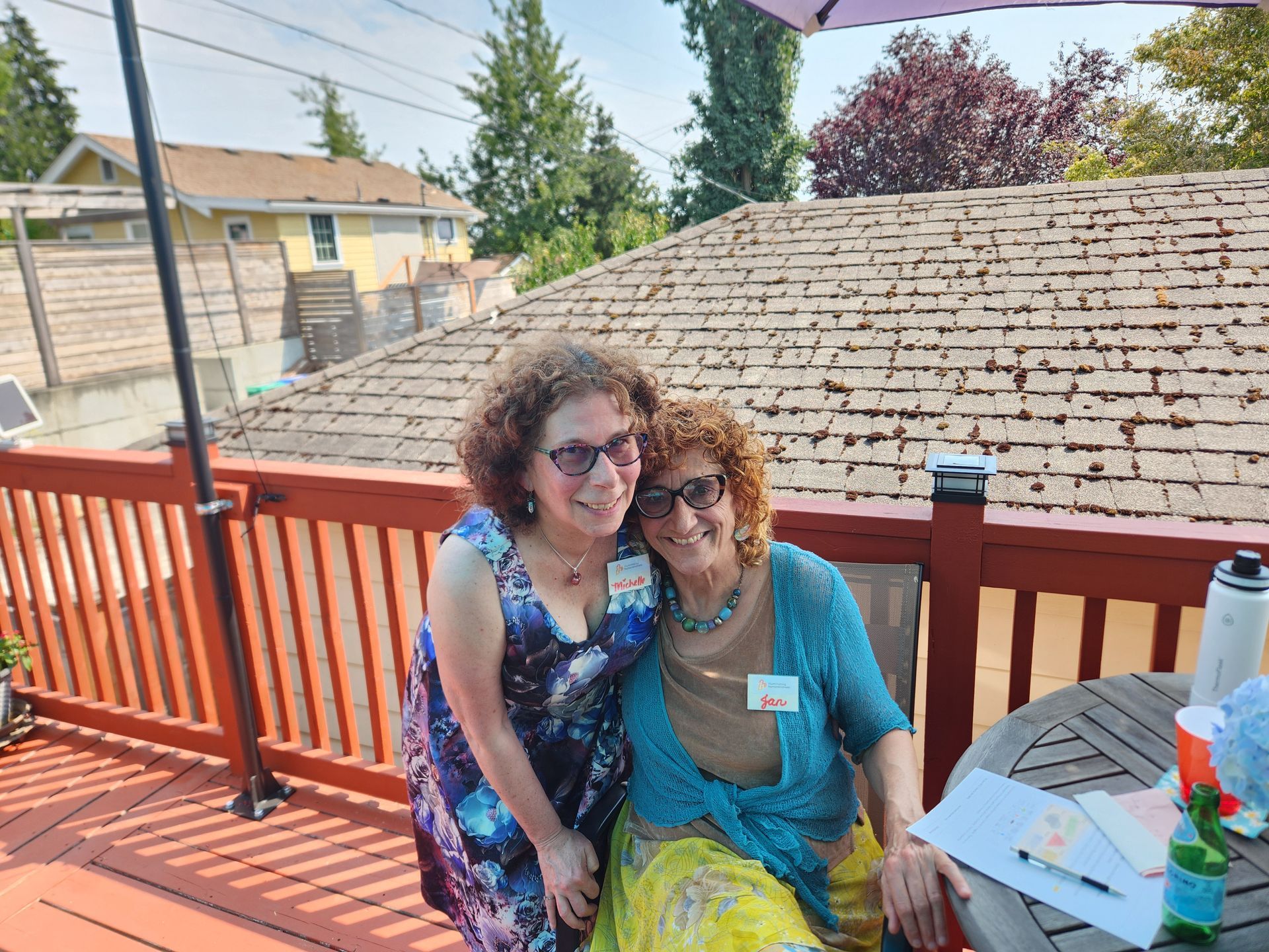 Two women smiling on a sunny deck. One in a blue dress, the other in a turquoise top and yellow skirt, under a sun umbrella.