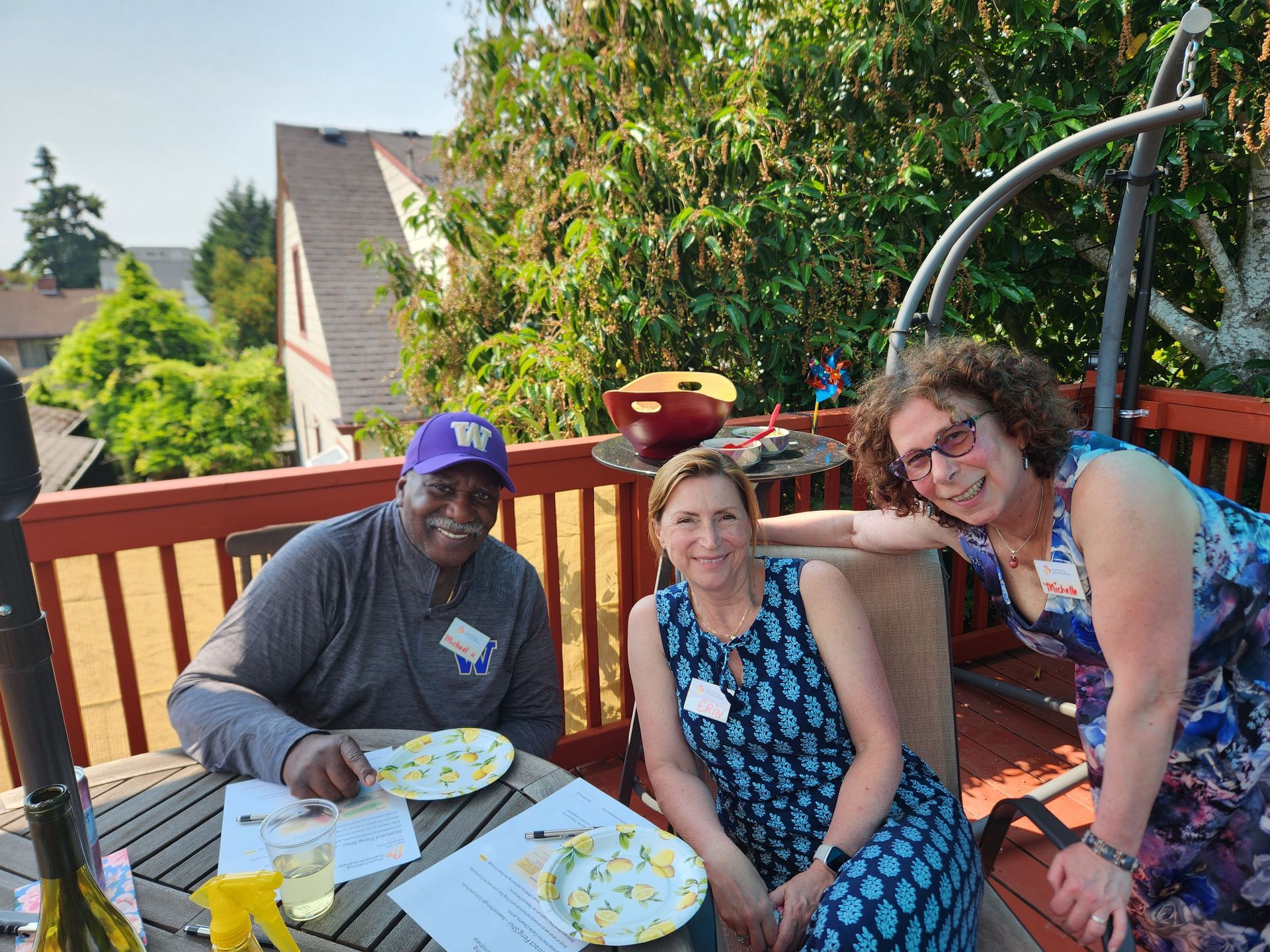 Three people at an outdoor party on a deck; smiles, sunny, plates, food, wine, lush trees in background.