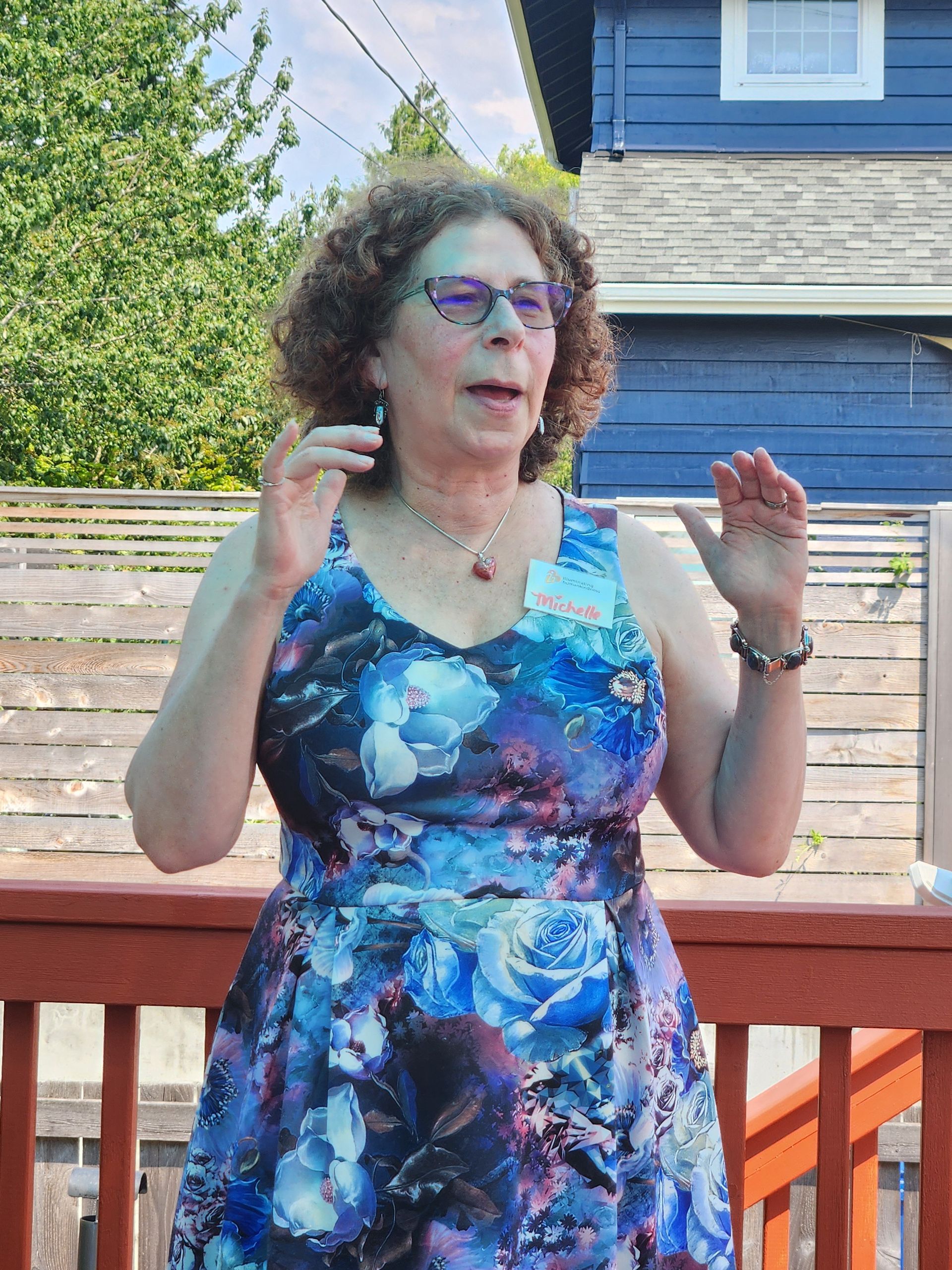 Woman in floral dress with hands raised outdoors on deck, blue house in background.