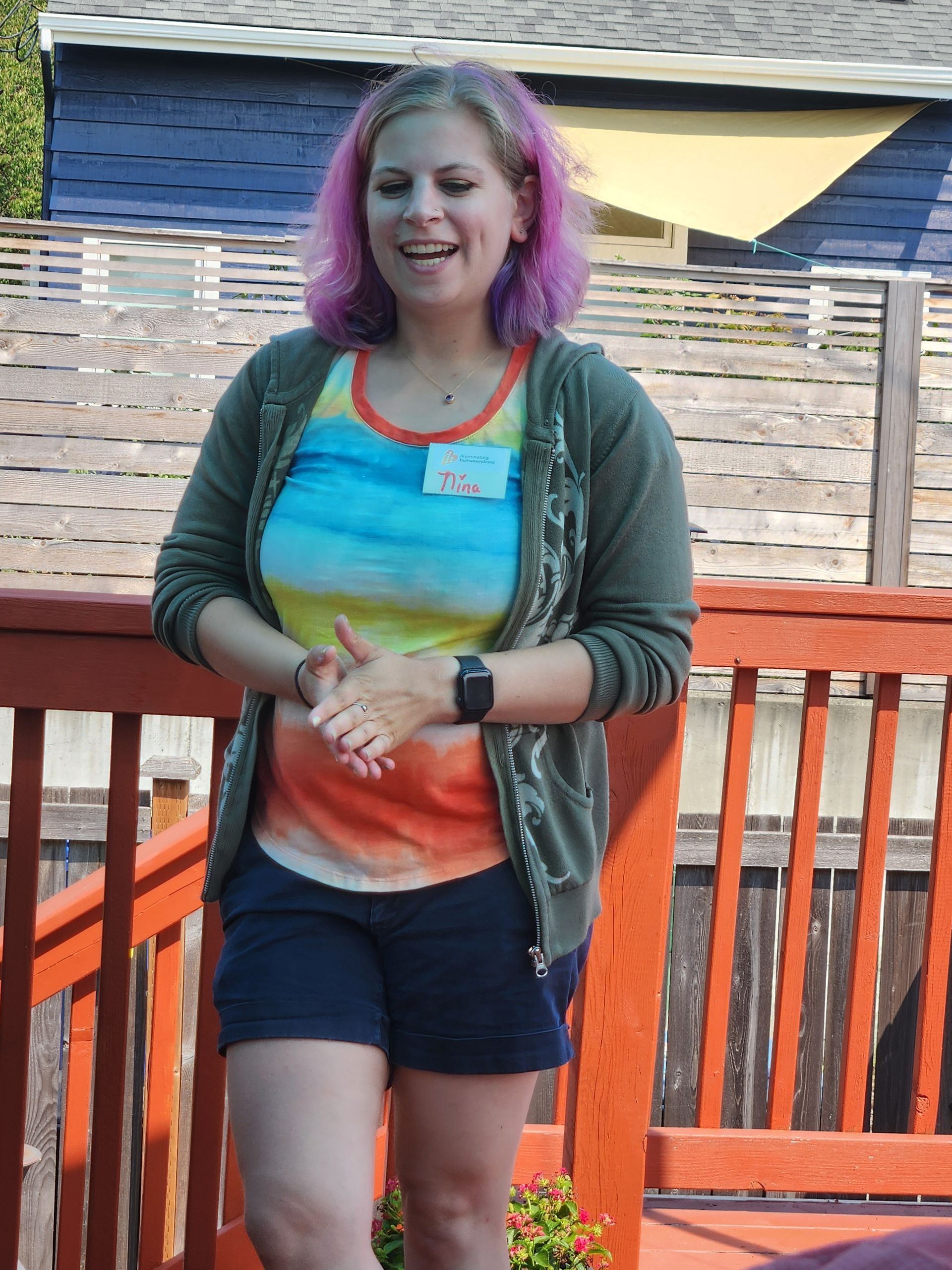 Woman with pink hair speaking on a wooden deck, wearing a colorful shirt and shorts.