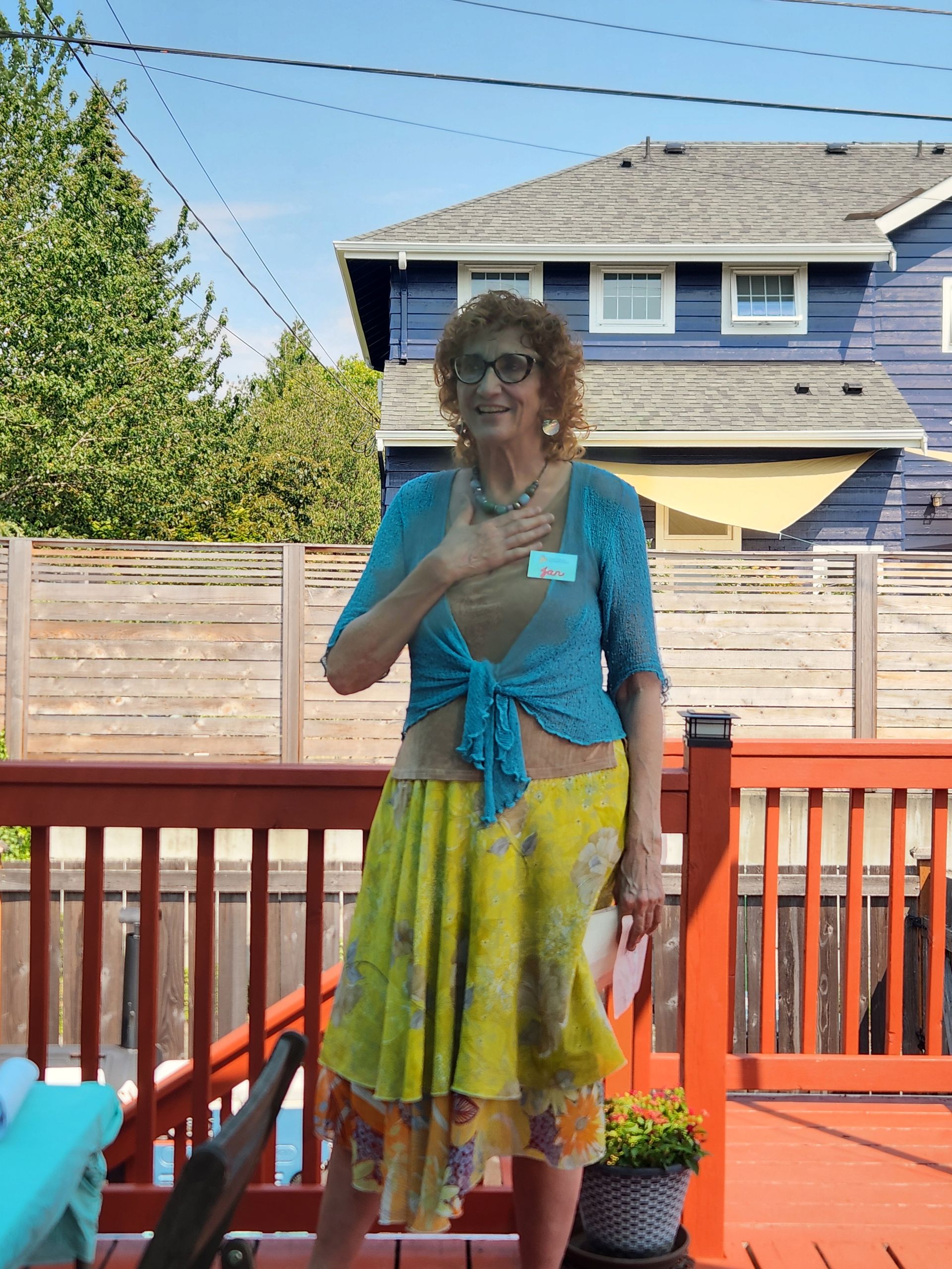 Woman on a deck wearing a blue top and yellow skirt, hand on her chest, with a blue house in the background.
