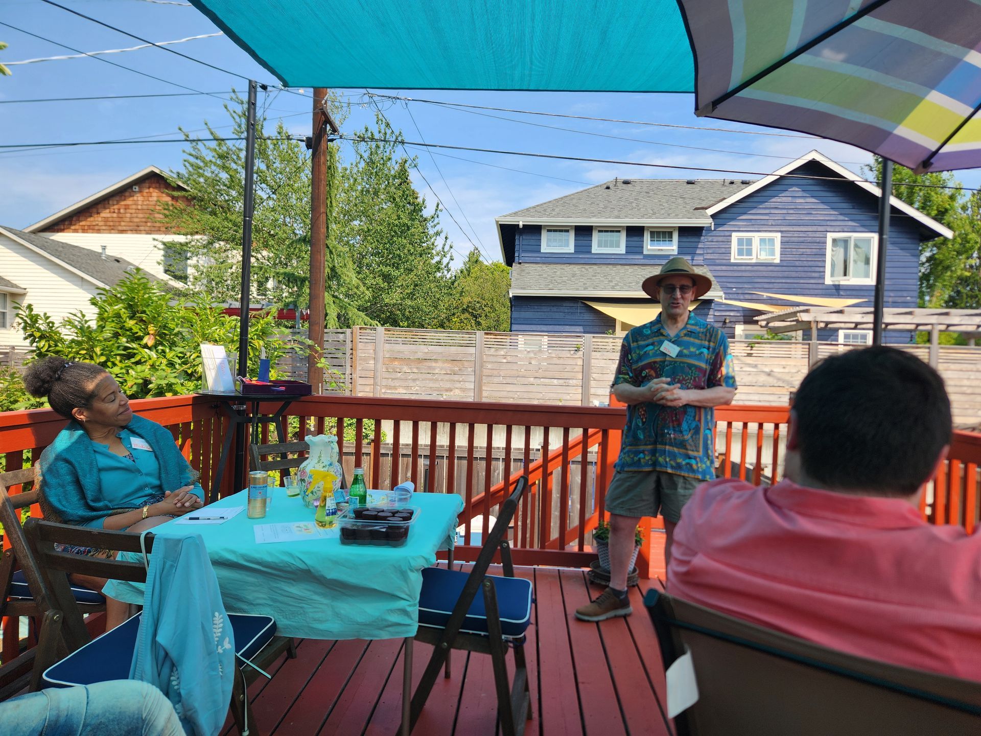 People on a deck, some seated at a table, one person speaking. Bright blue awning overhead, houses in the background.