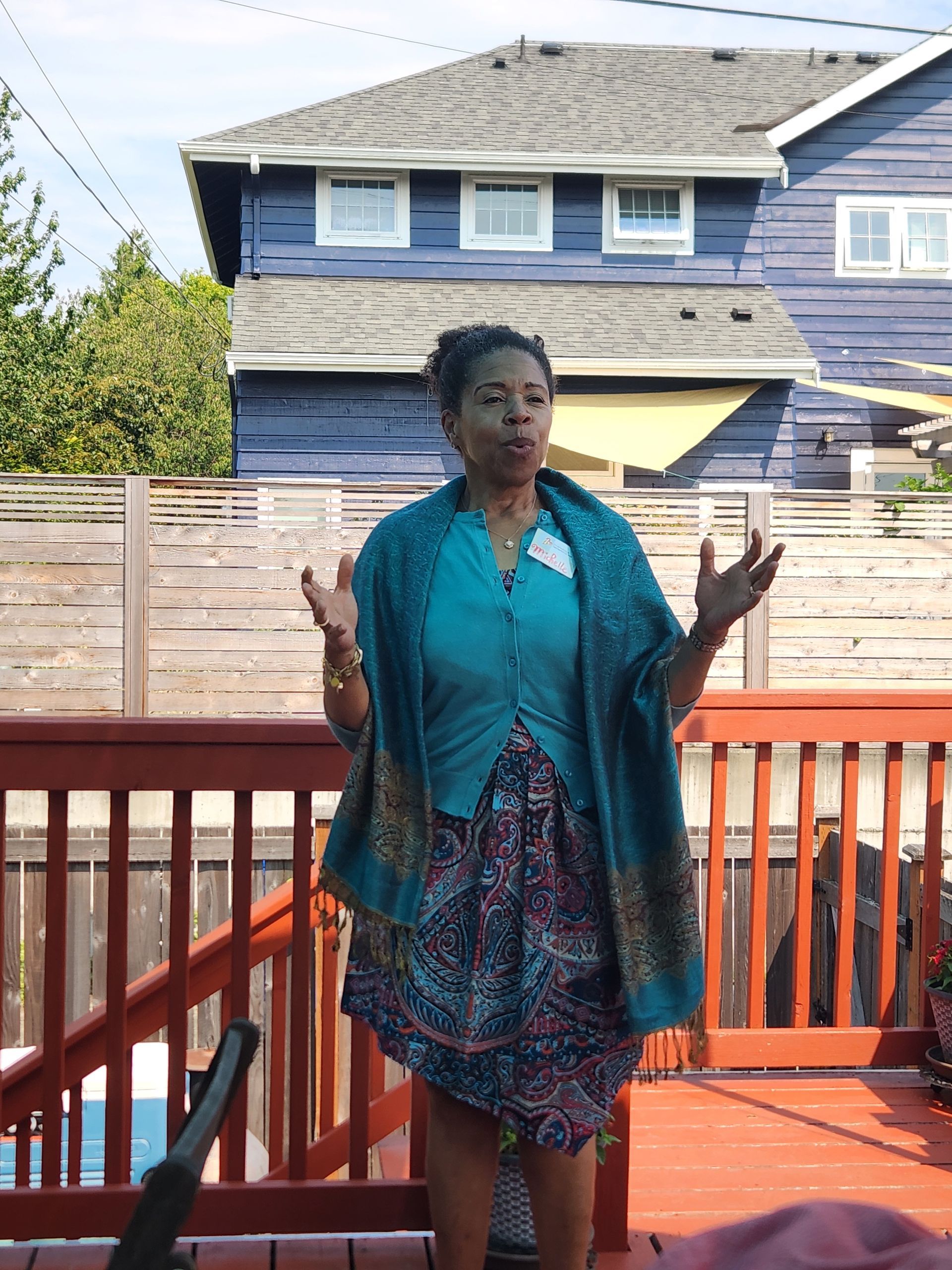 Woman speaking on a wooden deck, wearing teal shawl and patterned dress, with blue house and fence in background.