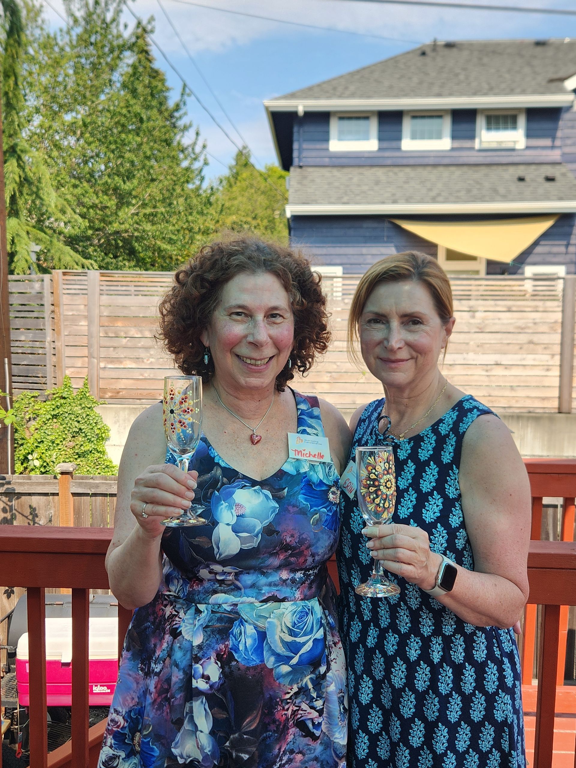 Two women toasting with champagne glasses on a wooden deck. One wears a floral dress; the other, a blue patterned dress.
