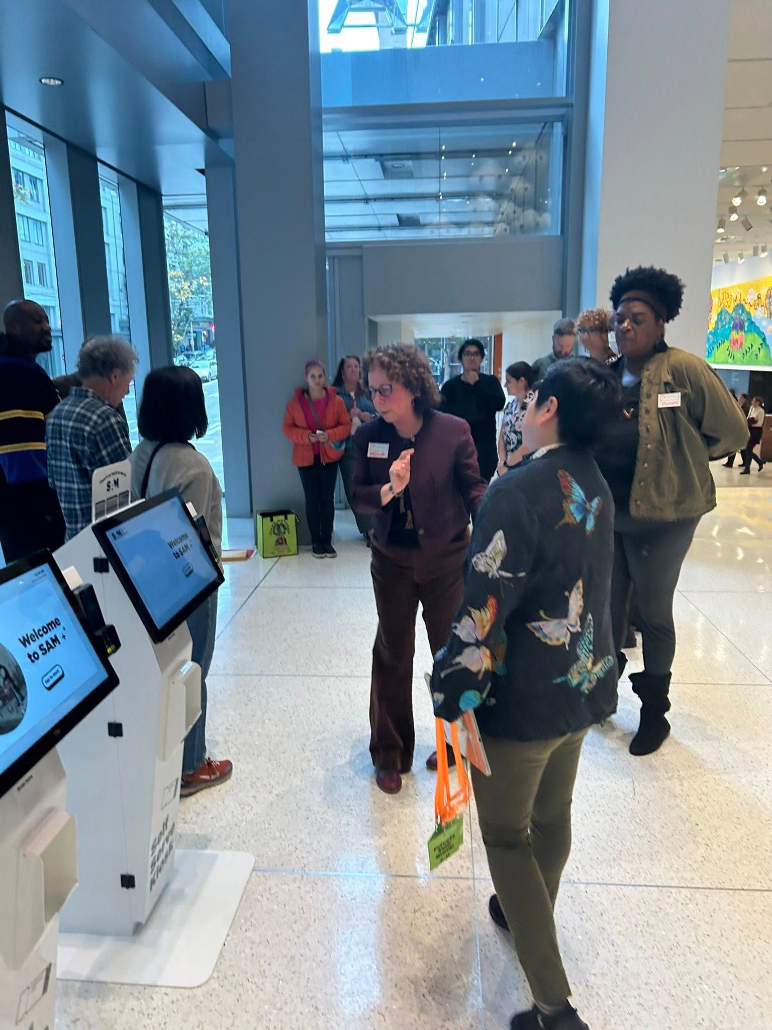 People in a modern building with kiosks. A group stands talking. Others look at kiosk screens.