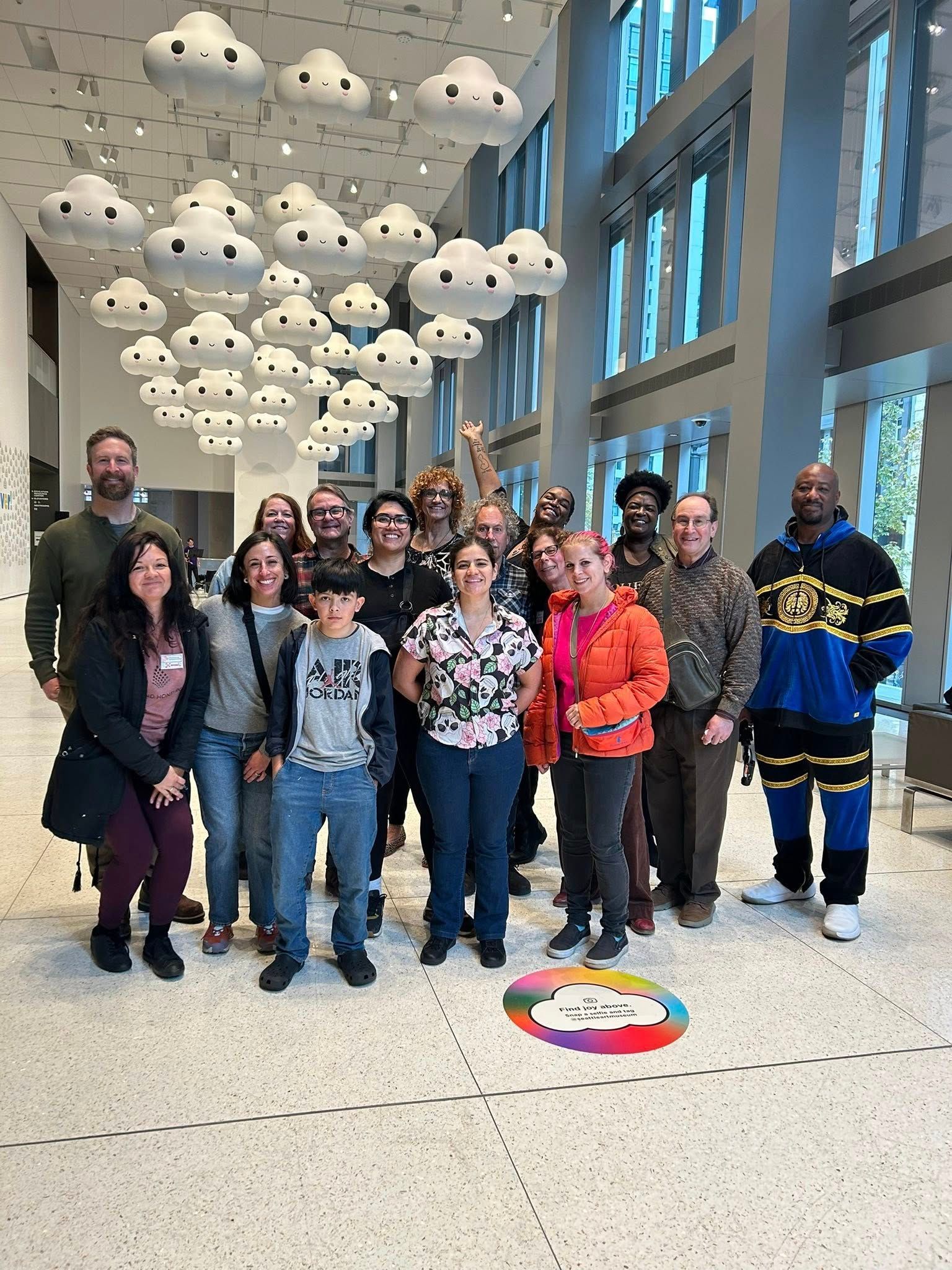 Group of people smiling, standing indoors under cloud art; large windows in background.