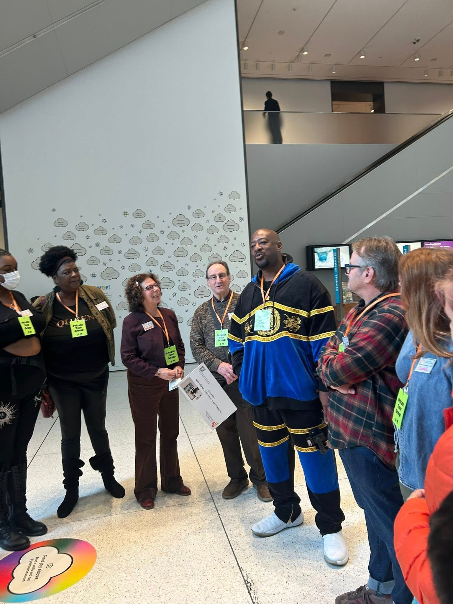 Group of people in a museum; some wearing name tags. Man in blue and gold jacket speaks. Modern building interior.