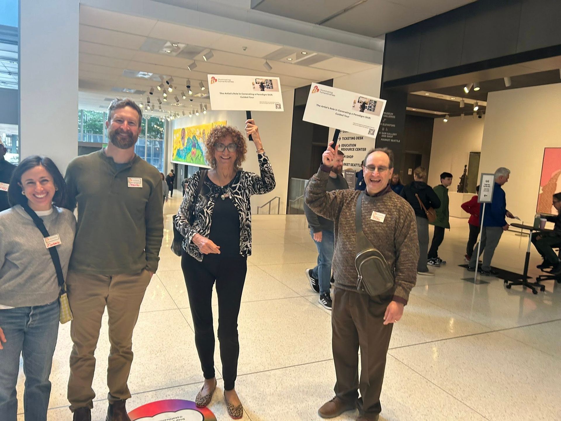 Four people hold signs in a museum hallway, smiling. Light-colored walls, artwork in the background.
