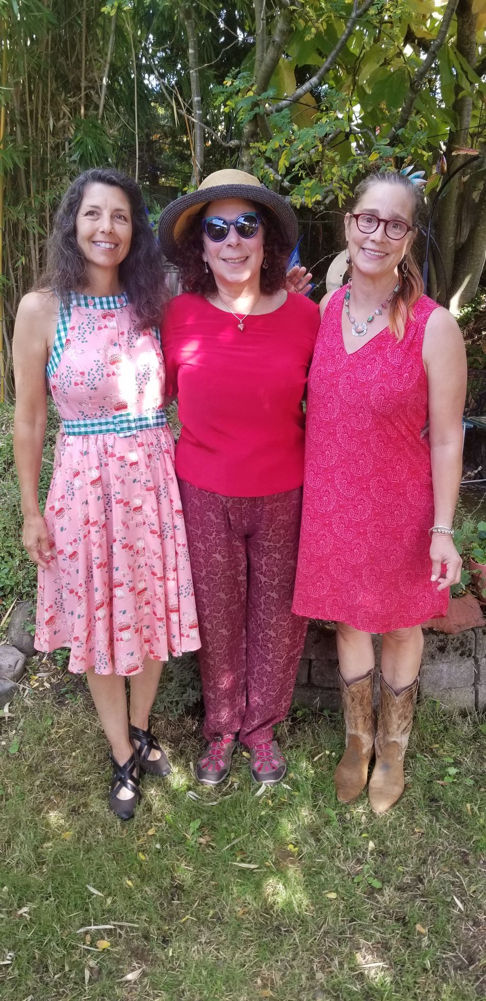 Three women standing outdoors, embracing. They wear dresses and pants in shades of red and pink.