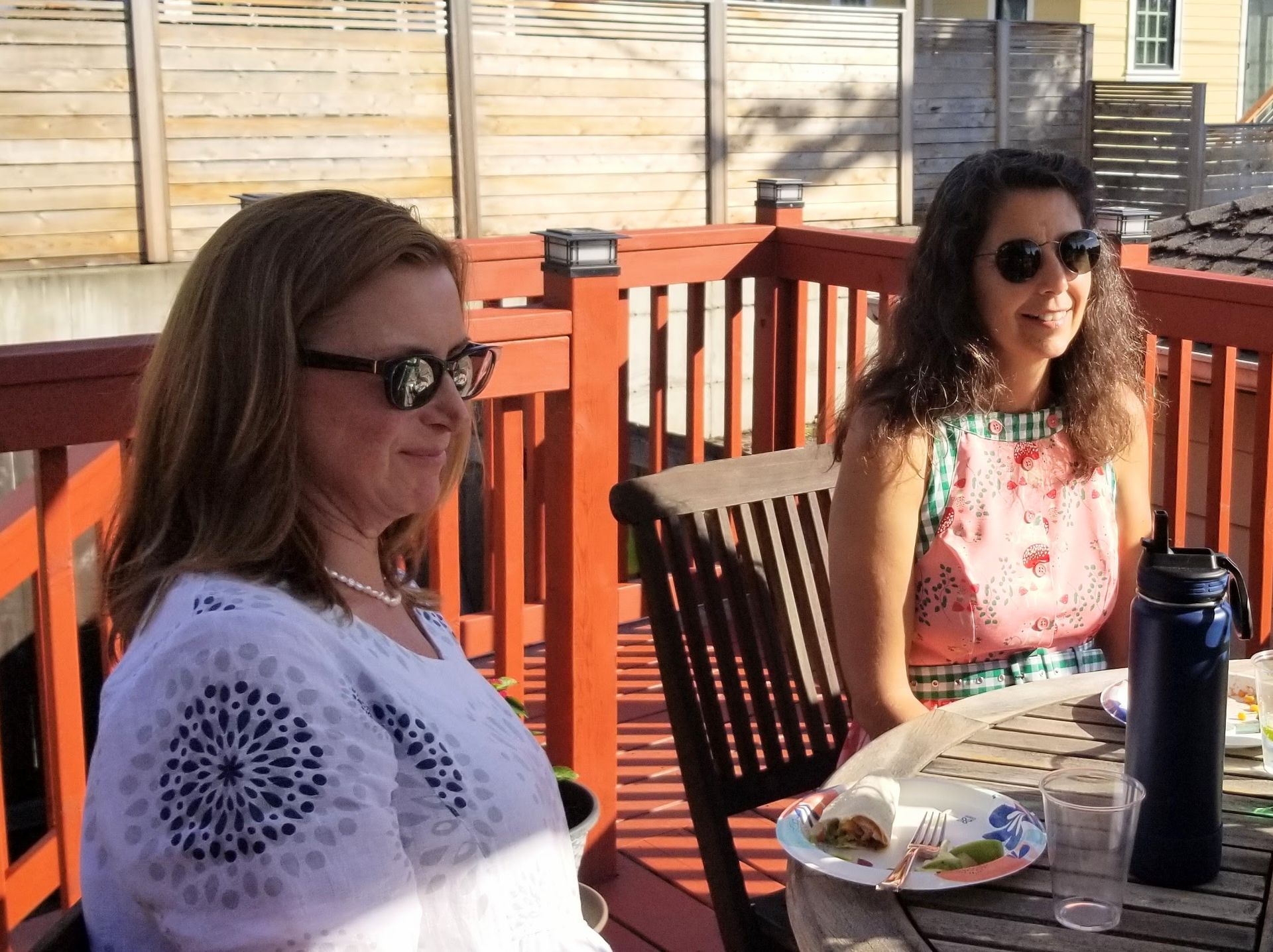 Two women wearing sunglasses sit at a wooden table on a deck; food, drinks, and a wooden fence are visible.