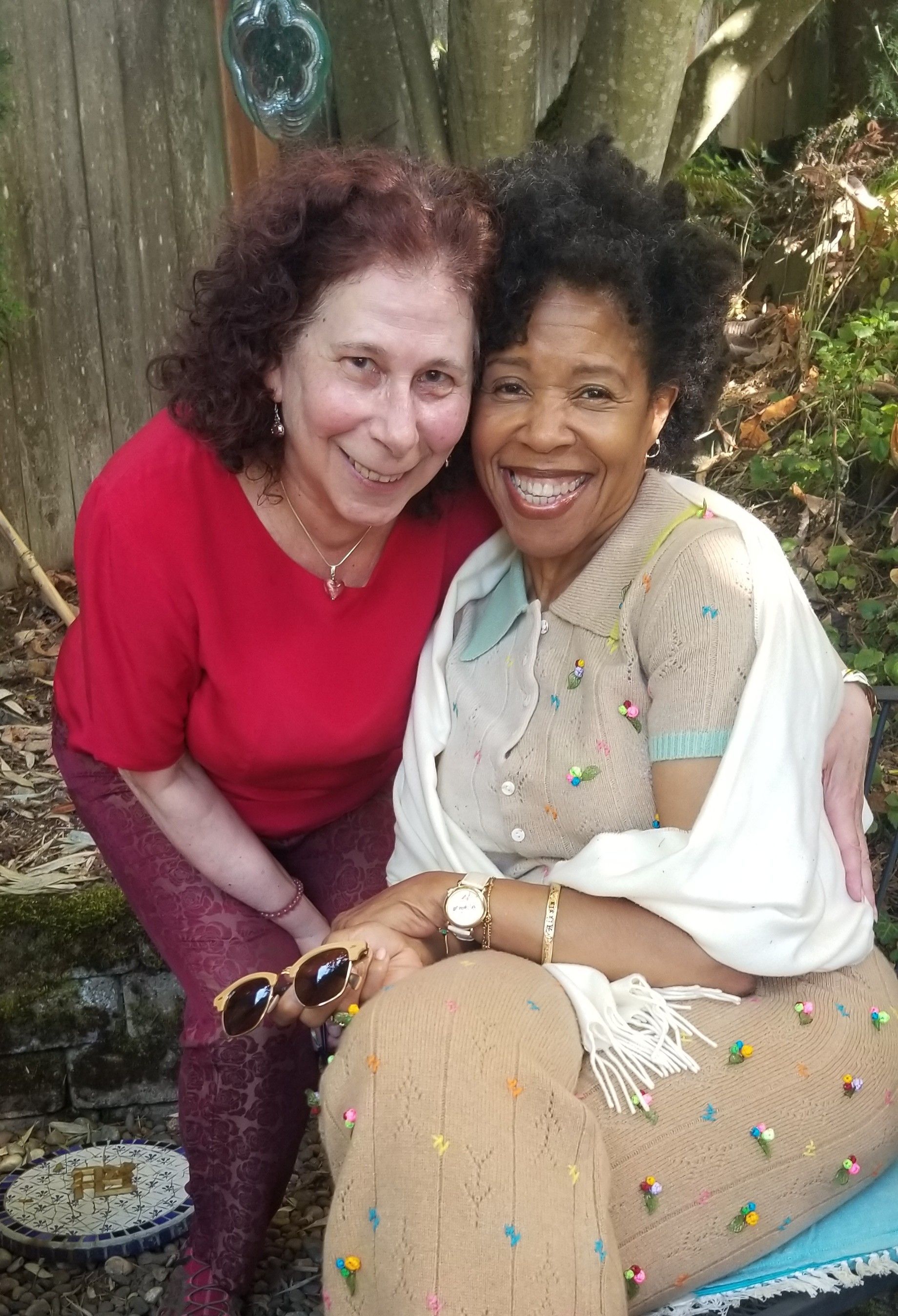 Two smiling women embracing outdoors. One in red, the other in beige with a white shawl.