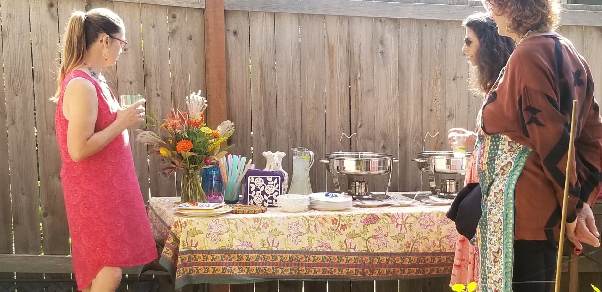 People at an outdoor gathering, buffet table with food and flowers, wooden fence background.