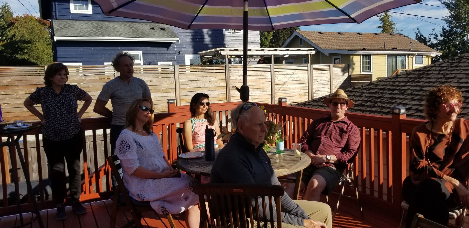 People gather on a deck under a striped umbrella. Houses are in the background.