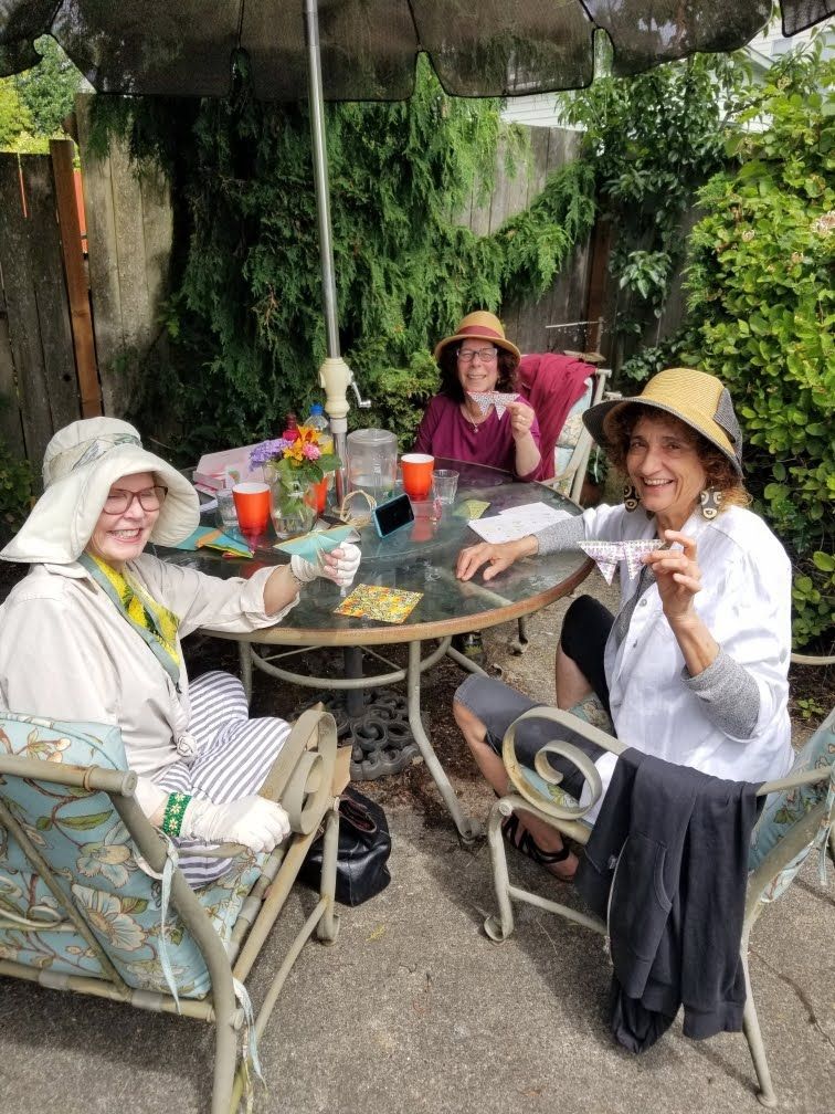 Three people in hats sit around a table outdoors, smiling, holding drinks and snacks, under an umbrella.