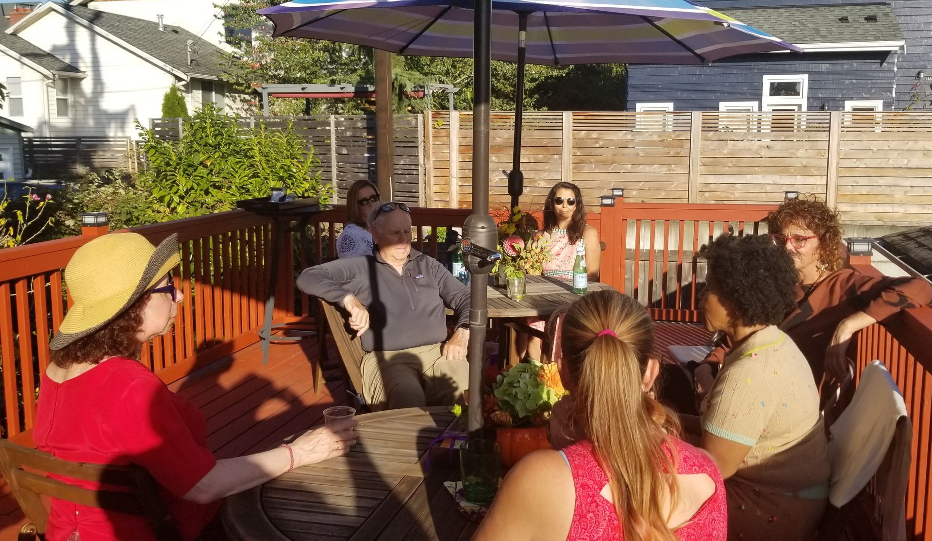 Group of people on a wooden deck under an umbrella, socializing and seated around a table.