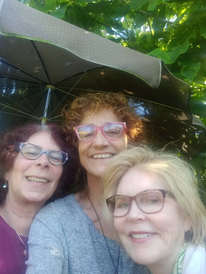 Three women smile for a selfie under a partially open umbrella, with foliage in the background.