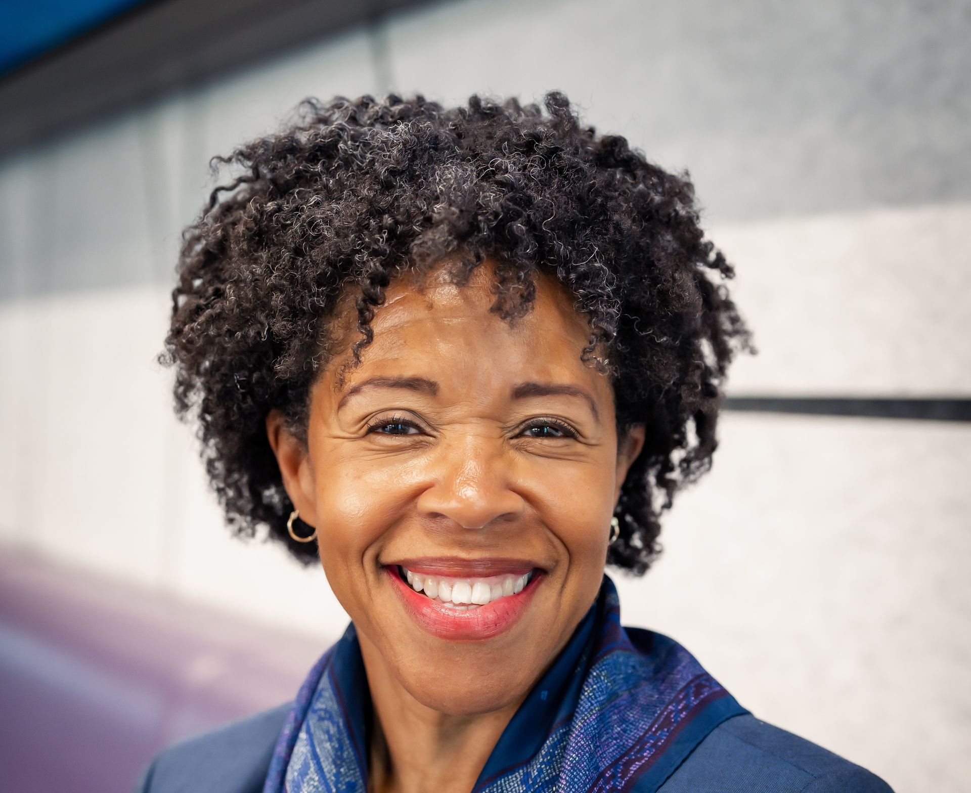Woman smiling, wearing a blue scarf and blazer, with short, curly dark hair.