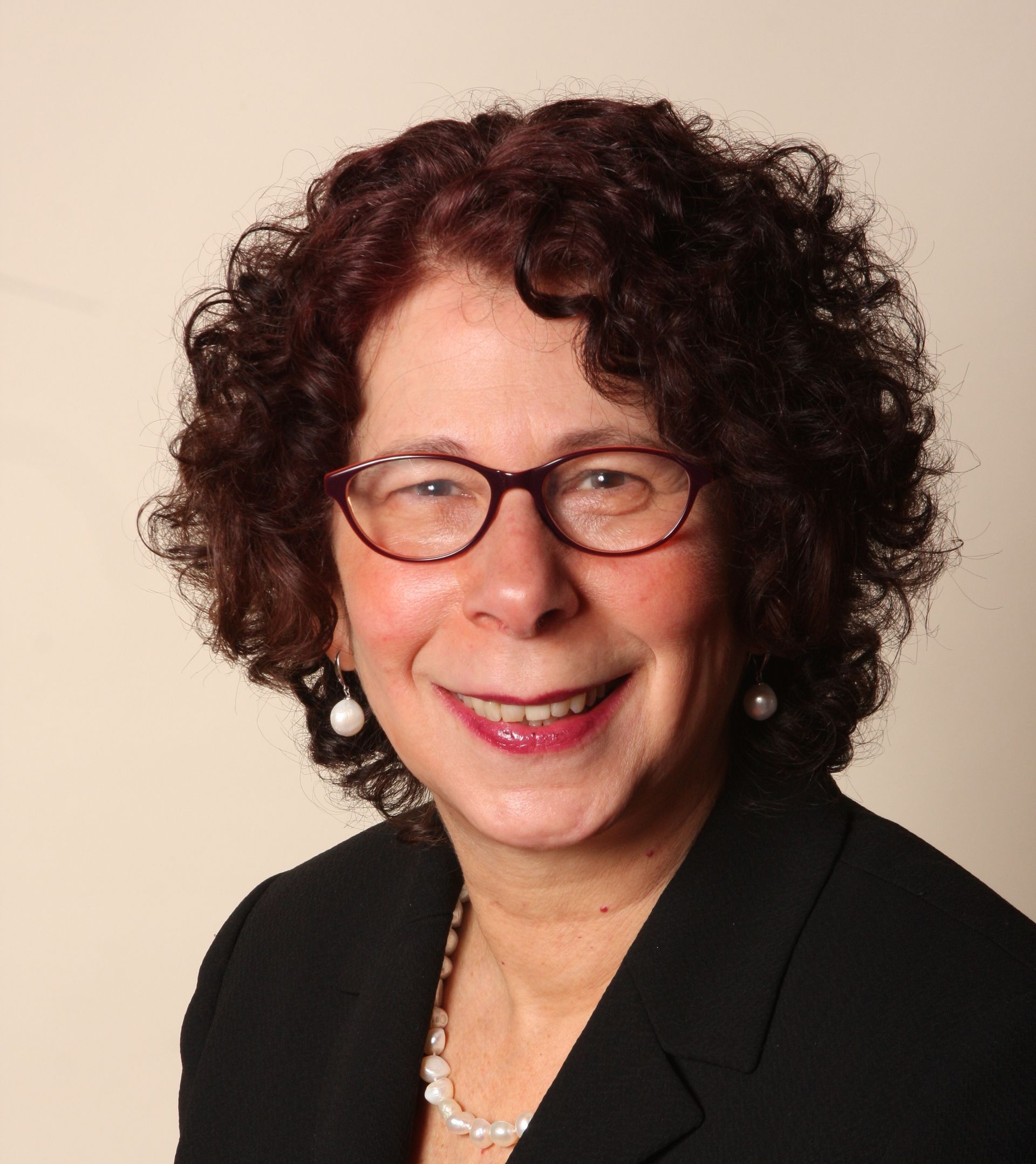 Woman with curly brown hair, glasses, and pearl necklace, smiling; wearing a black blazer.