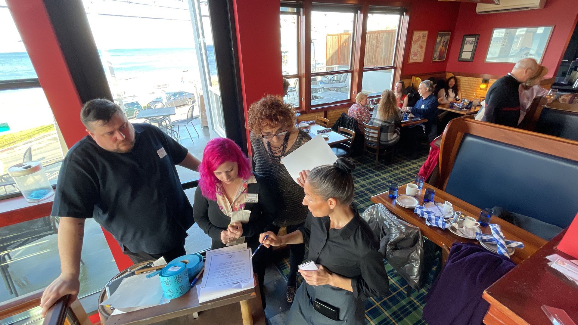 People gathered inside a restaurant with ocean views, reviewing papers and interacting with staff.