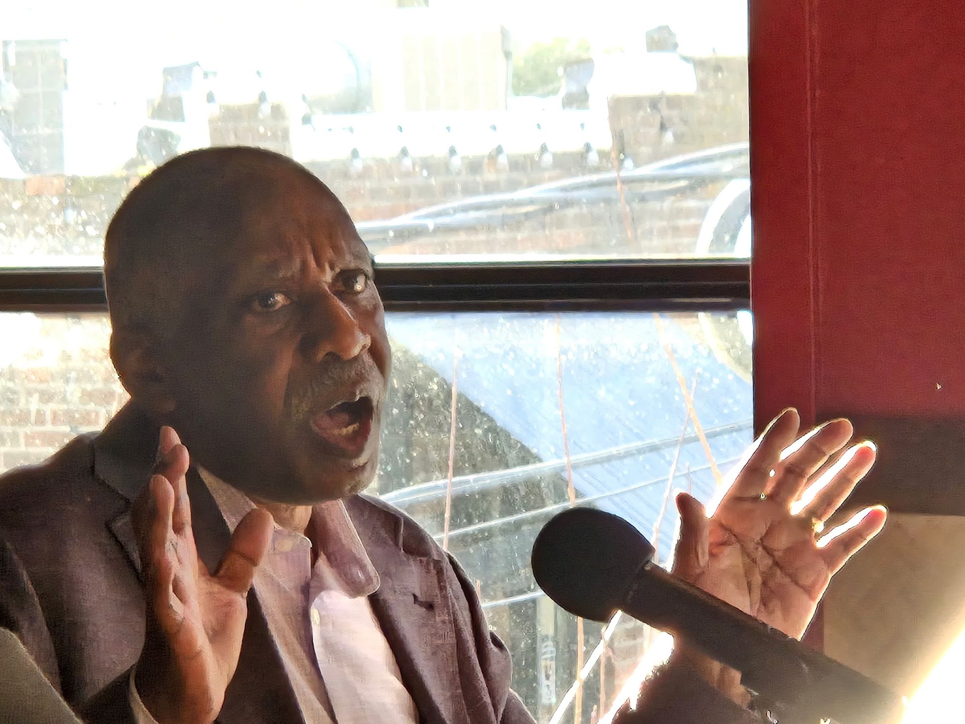 Man speaking into a microphone, gesturing with both hands. Indoors, against a window.