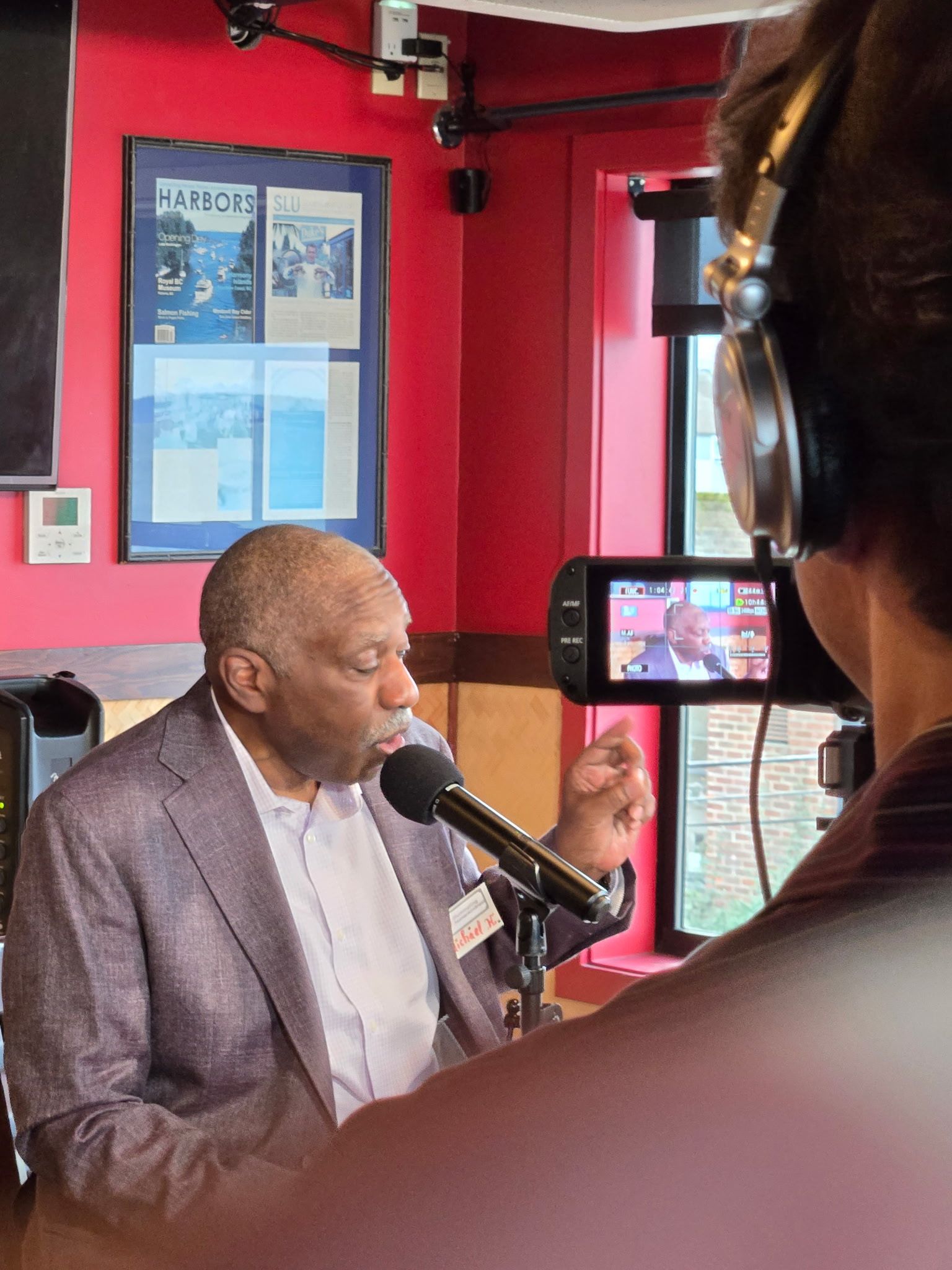 Man in suit speaks into microphone, being filmed. Red wall, framed print in background.