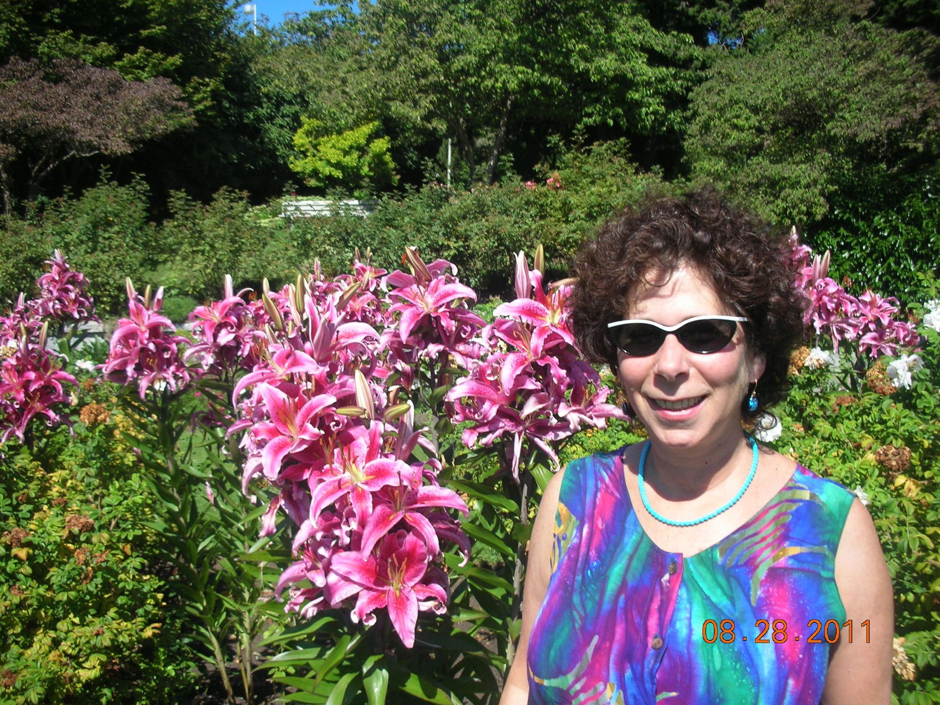 Woman in sunglasses smiles in front of pink lilies; sunny garden setting.