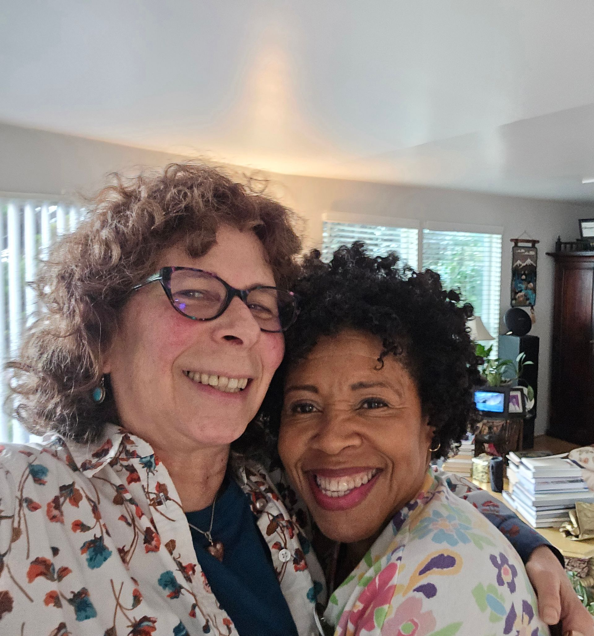 Two smiling women embrace; one with glasses and curly hair, the other with short dark hair, both wearing floral shirts.