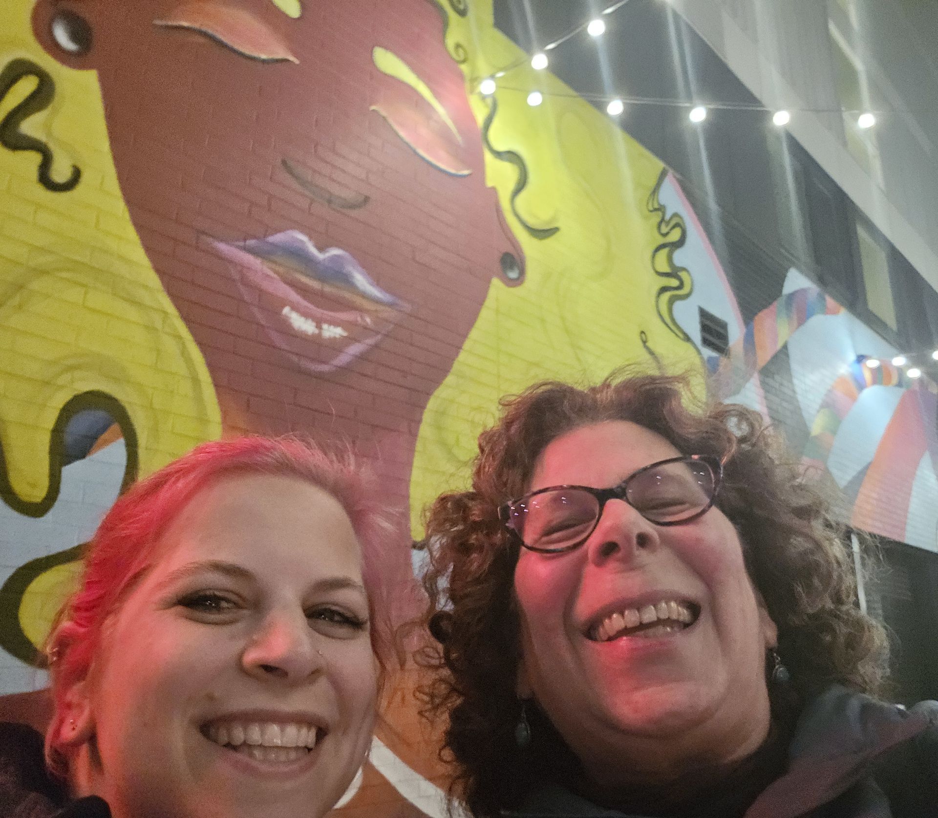 Two smiling people posing in front of a colorful mural of a woman's face.