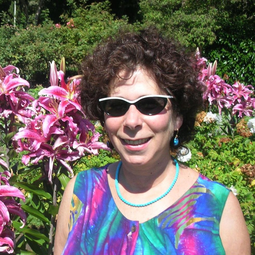 Woman wearing sunglasses smiles amidst pink lilies in a garden. Turquoise jewelry and colorful top.