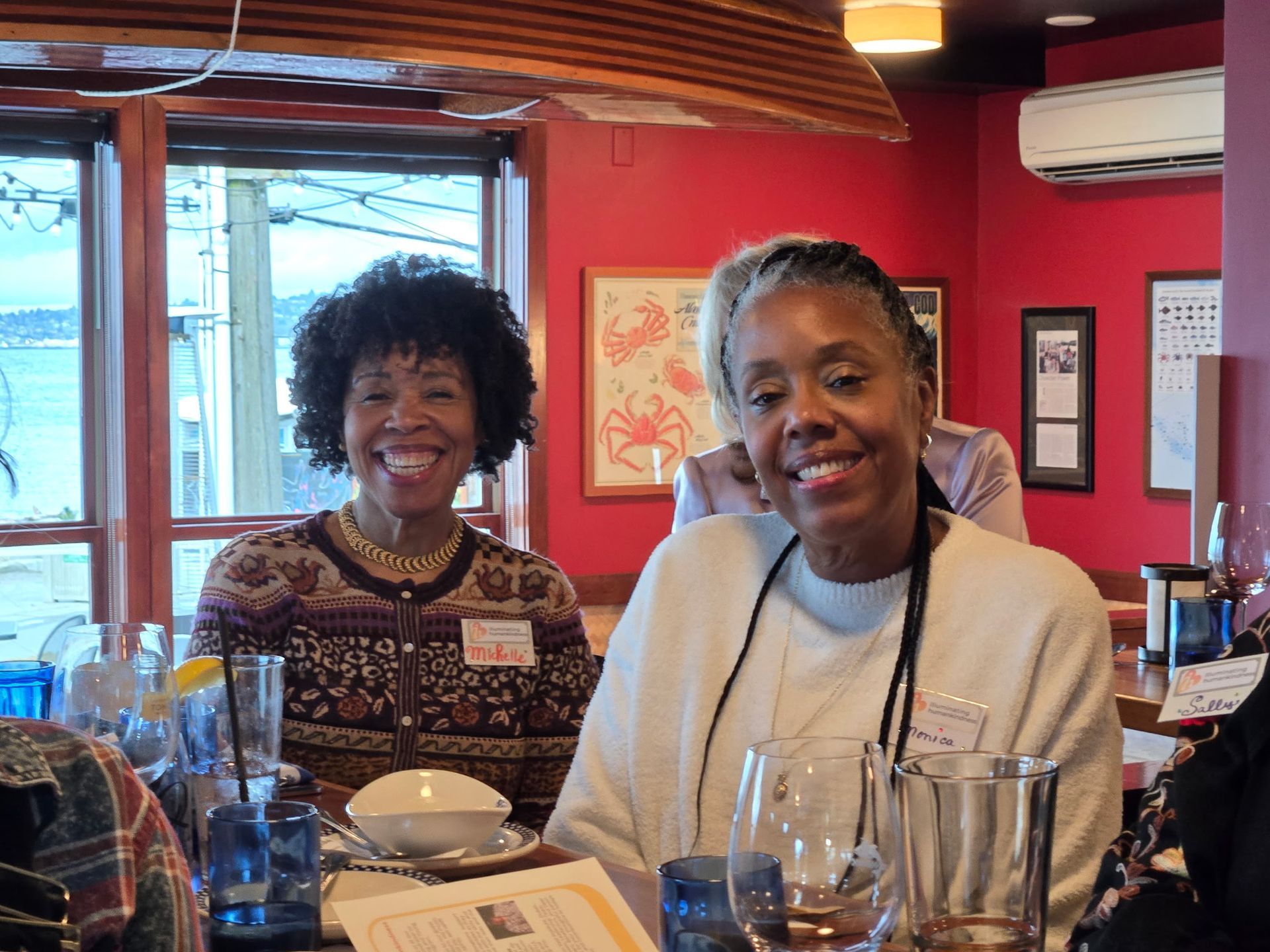 Two smiling women at a table in a restaurant with a red wall and a window view.