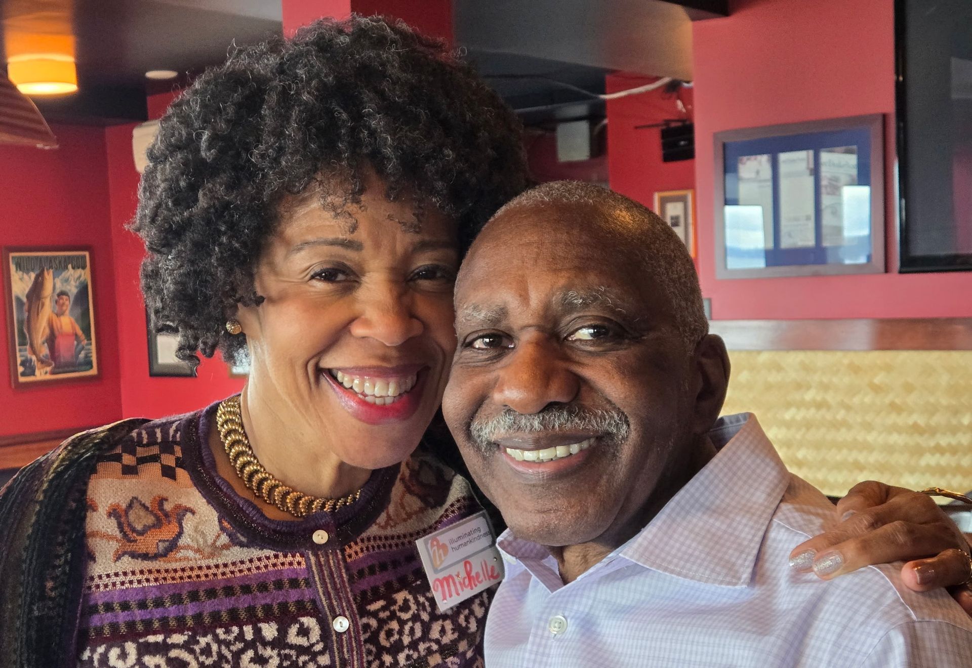 Two people smiling, indoors. Woman with curly hair, patterned top, and man with bald head, collared shirt.