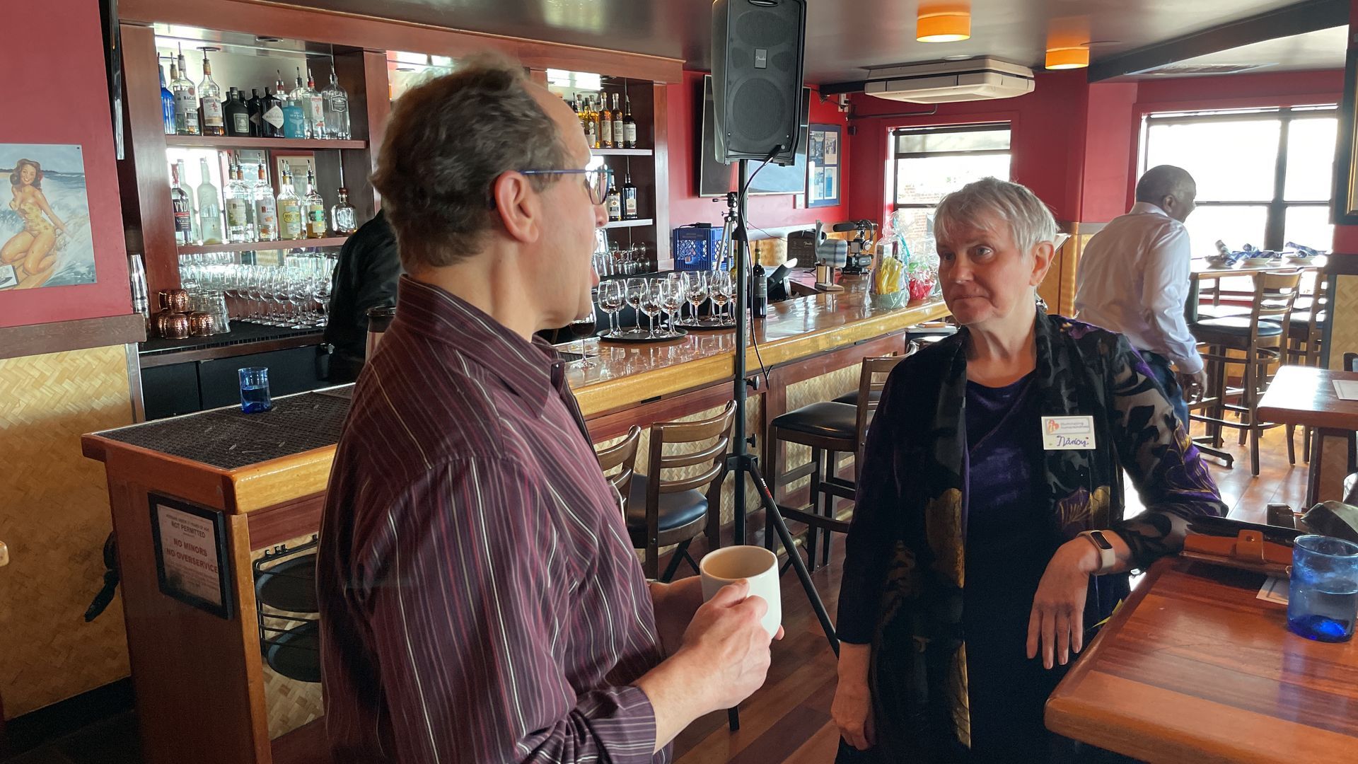 Two people talking at a bar; man holds a cup. The bar has bottles and a speaker.
