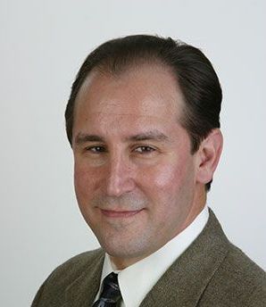 Man in suit smiling, facing camera; neutral expression, light background.
