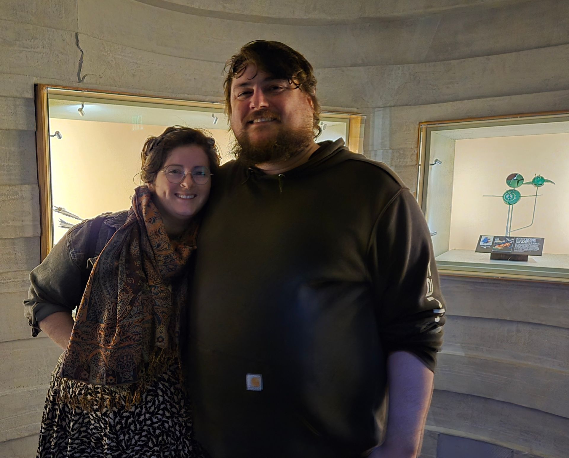 Two people smiling, standing near display cases with art in a dimly lit room.