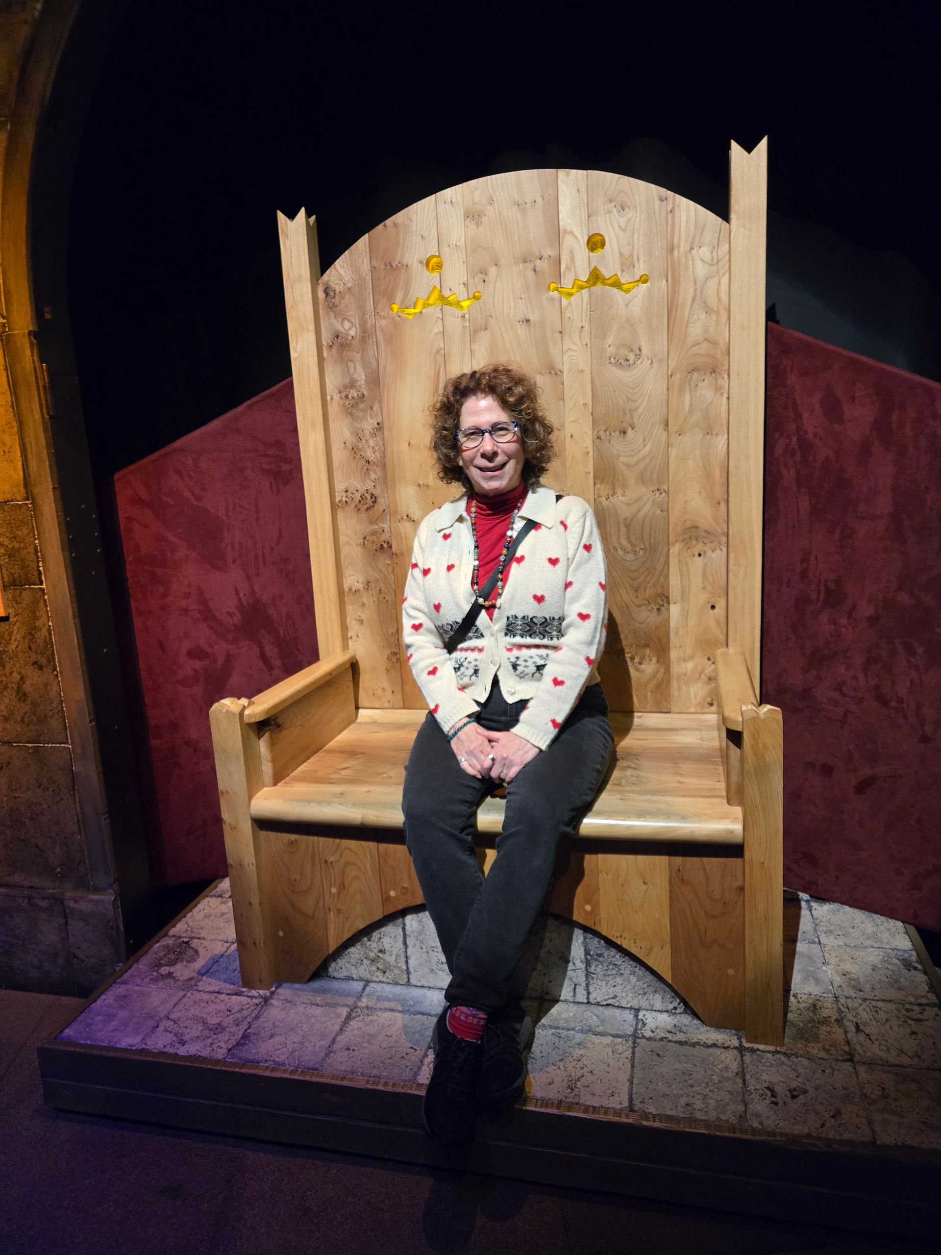 Woman sitting on a large wooden throne with a dark setting and a red backdrop.