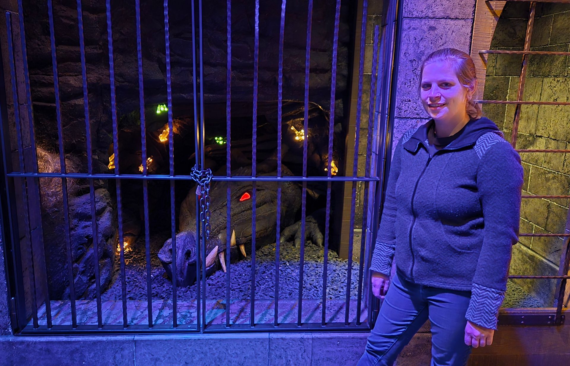 Woman stands near a cage, a dragon statue inside. Dark blue lighting.