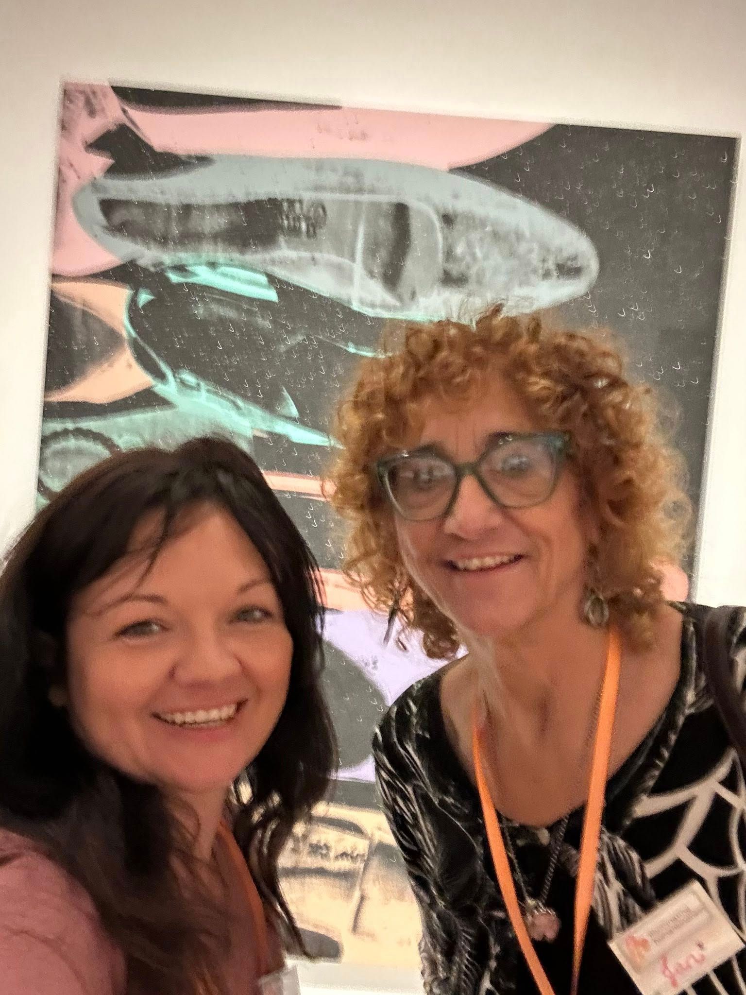 Two women smiling in front of a colorful abstract artwork of a plane.