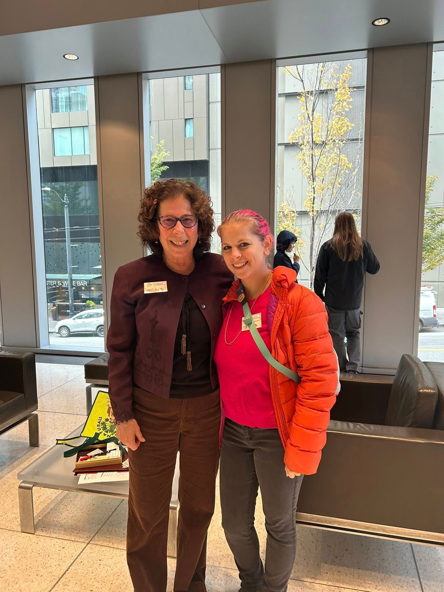 Two women pose together inside a building. One wears a purple jacket, the other an orange coat. Windows and an outdoor view are in the background.