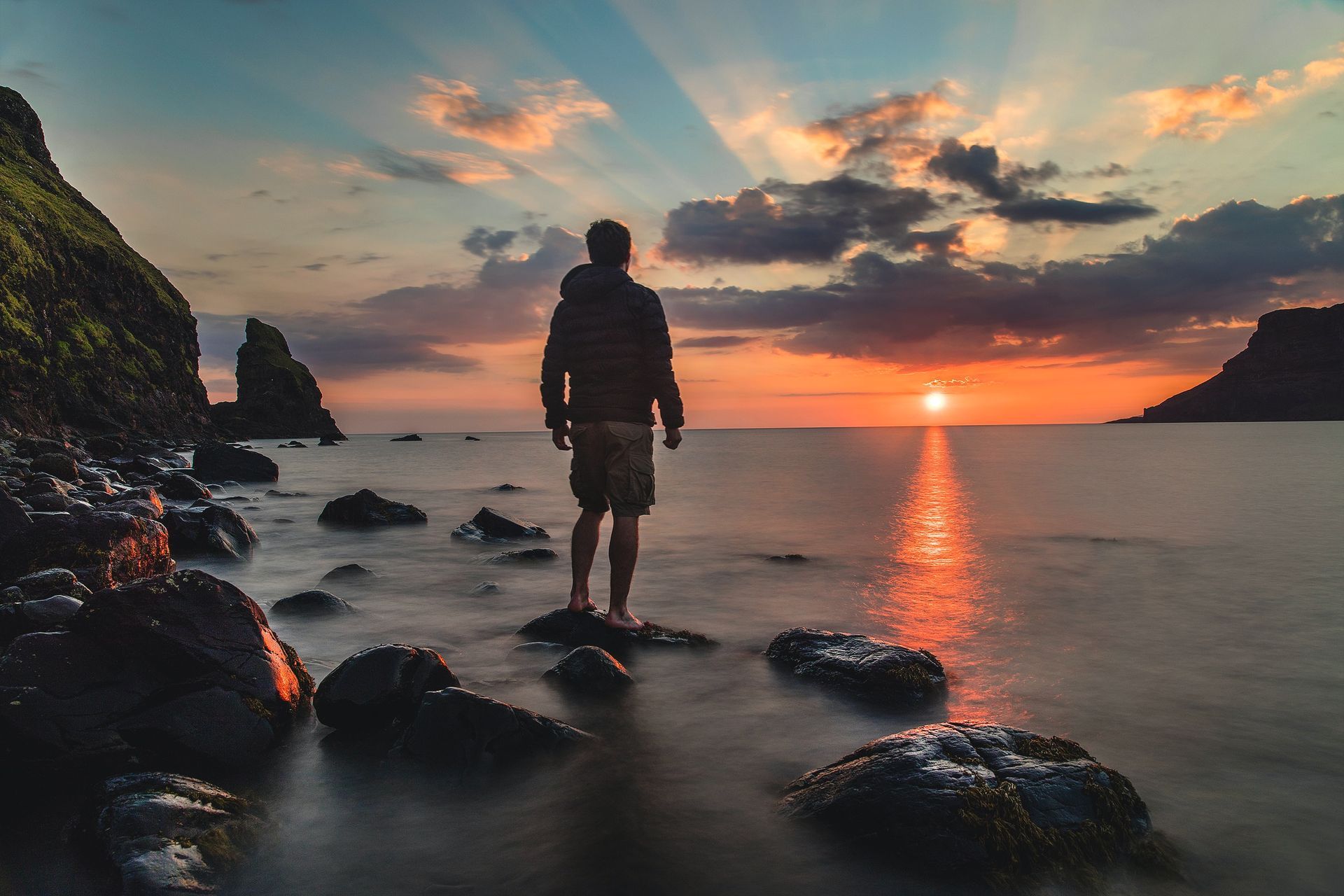 Man standing on rocks, facing a sunset over the ocean. Orange, blue sky.