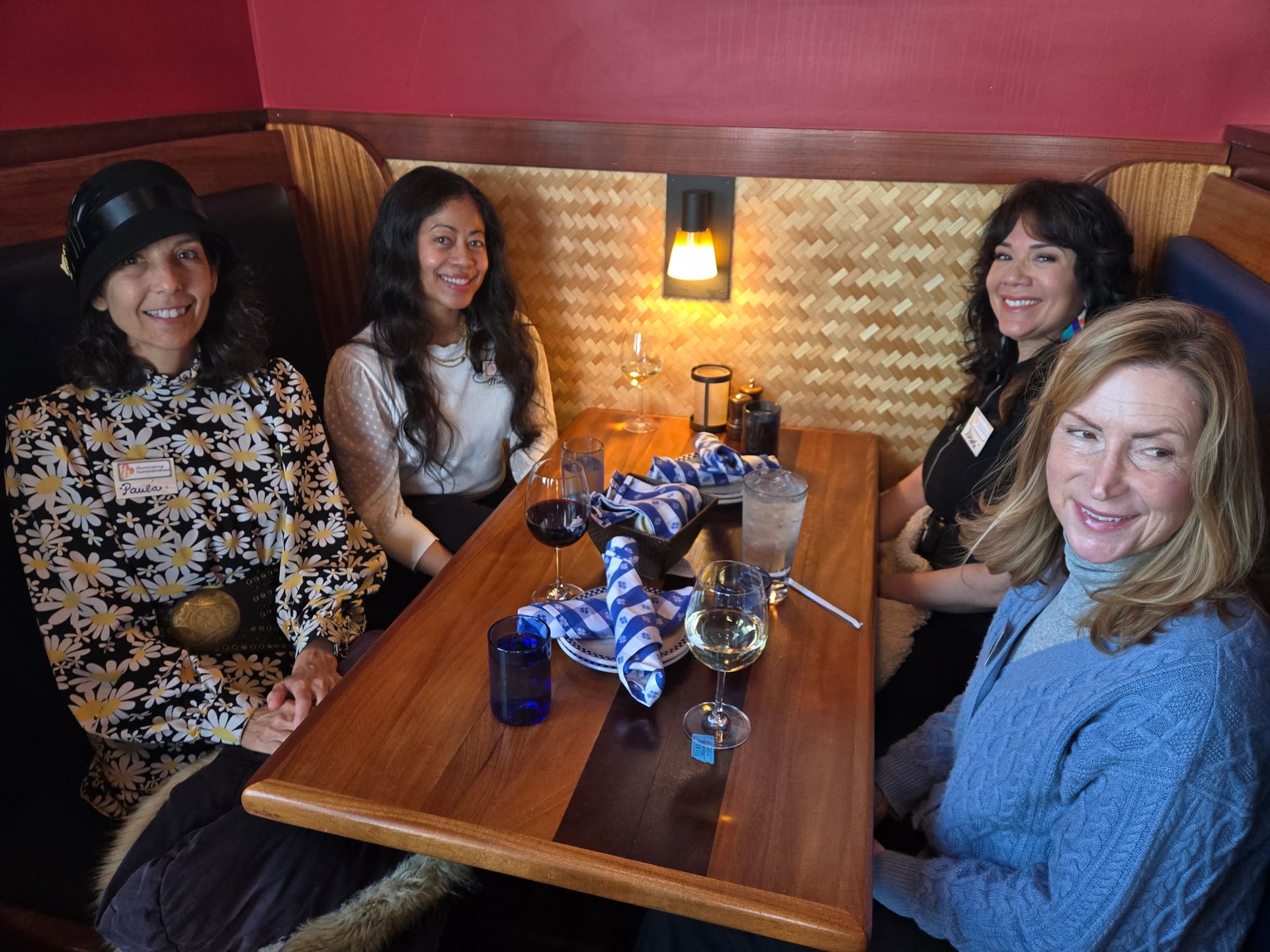 Four people seated at a table in a restaurant booth, smiling. There's food, drinks and a decorative setting.