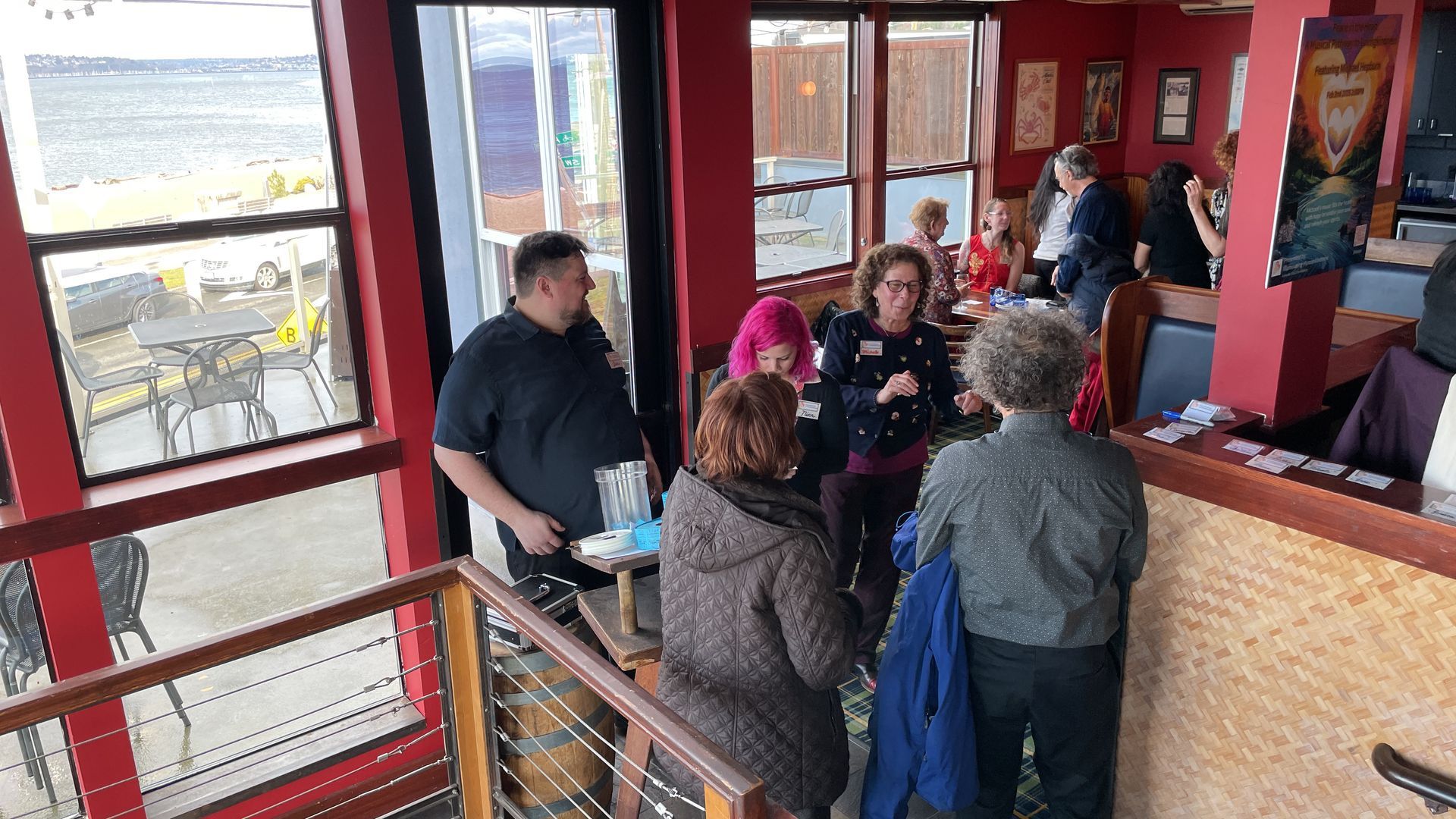 People gathering in a red-walled restaurant near windows overlooking water.