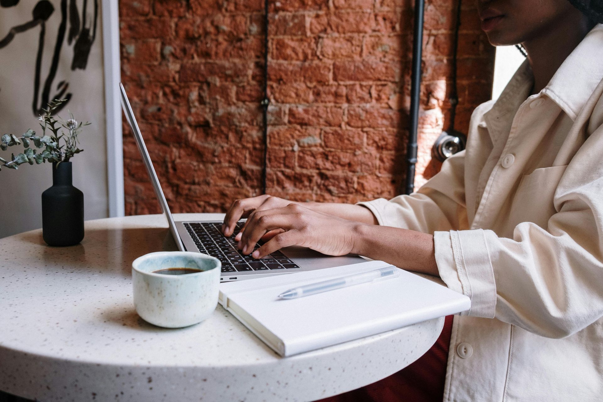 Person typing on laptop at a table, with a coffee cup and notebook. Brick wall in the background.